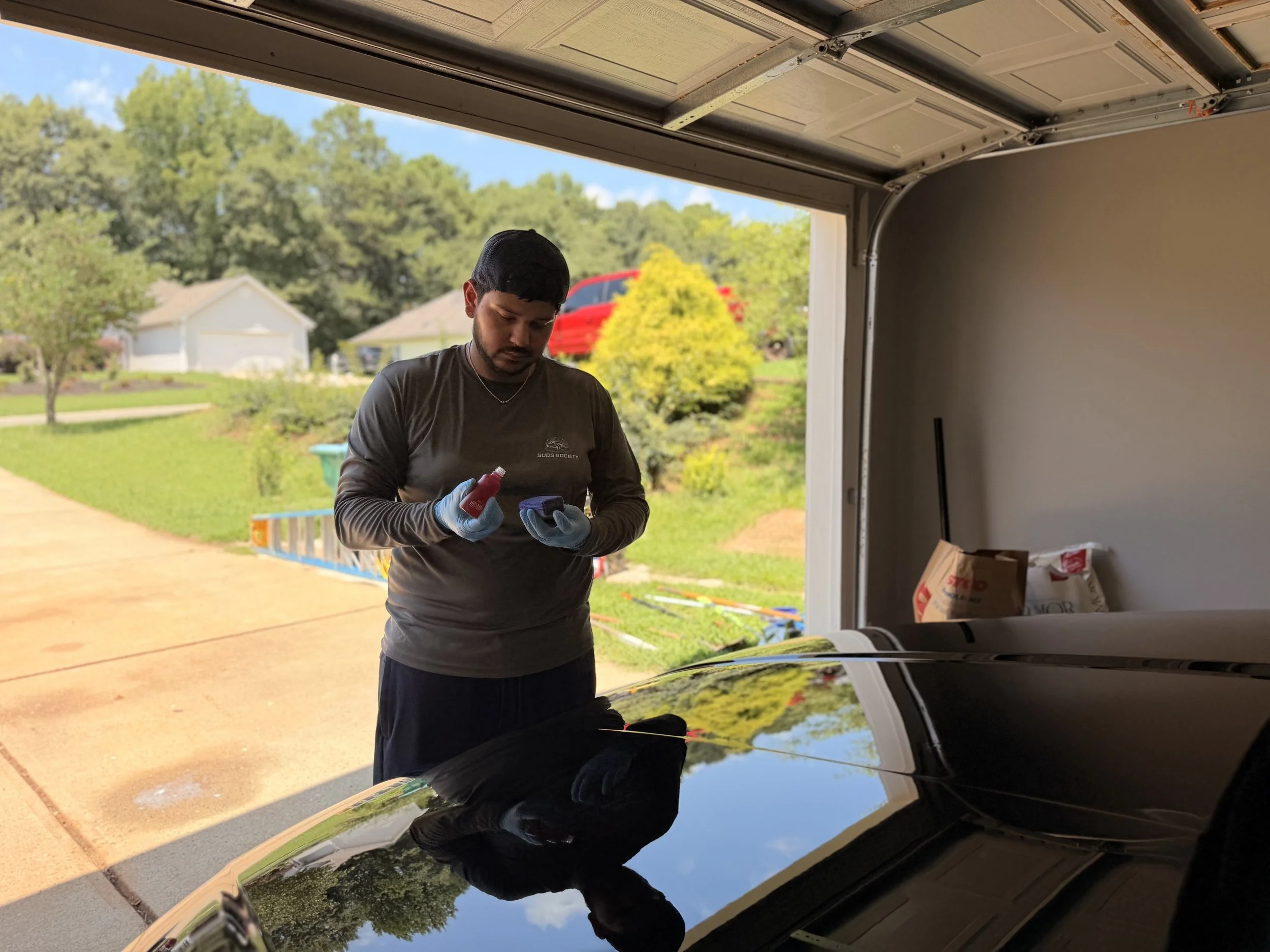 Man in a black shirt and black cap, wearing gloves, standing in a garage next to a car, looking at a small object in his hands, with a suburban neighborhood and trees in the background.