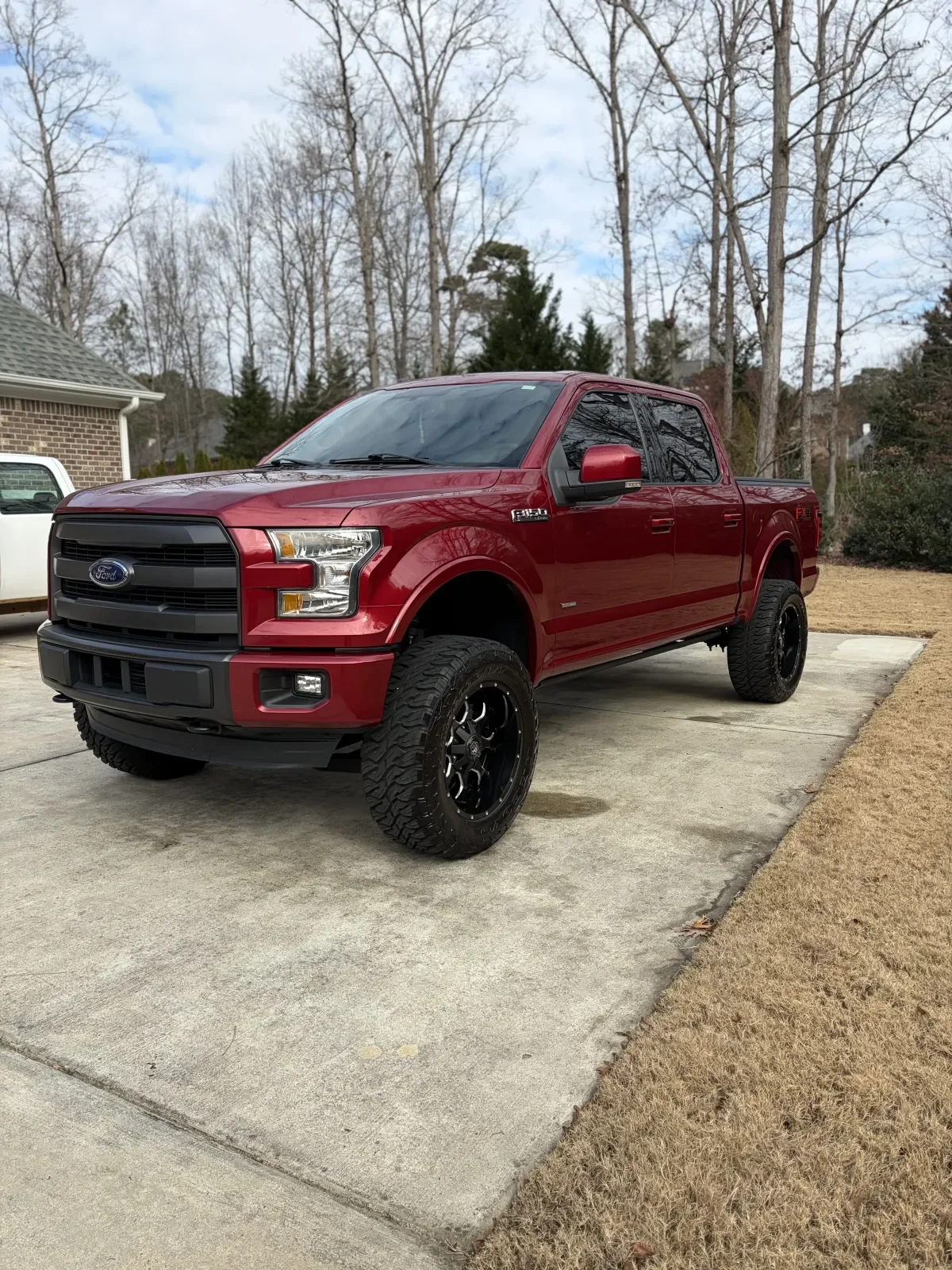 A red Ford F-150 pickup truck with black wheels parked on a concrete driveway in a suburban yard, with a grassy area, trees, a house, and cloudy sky in the background.