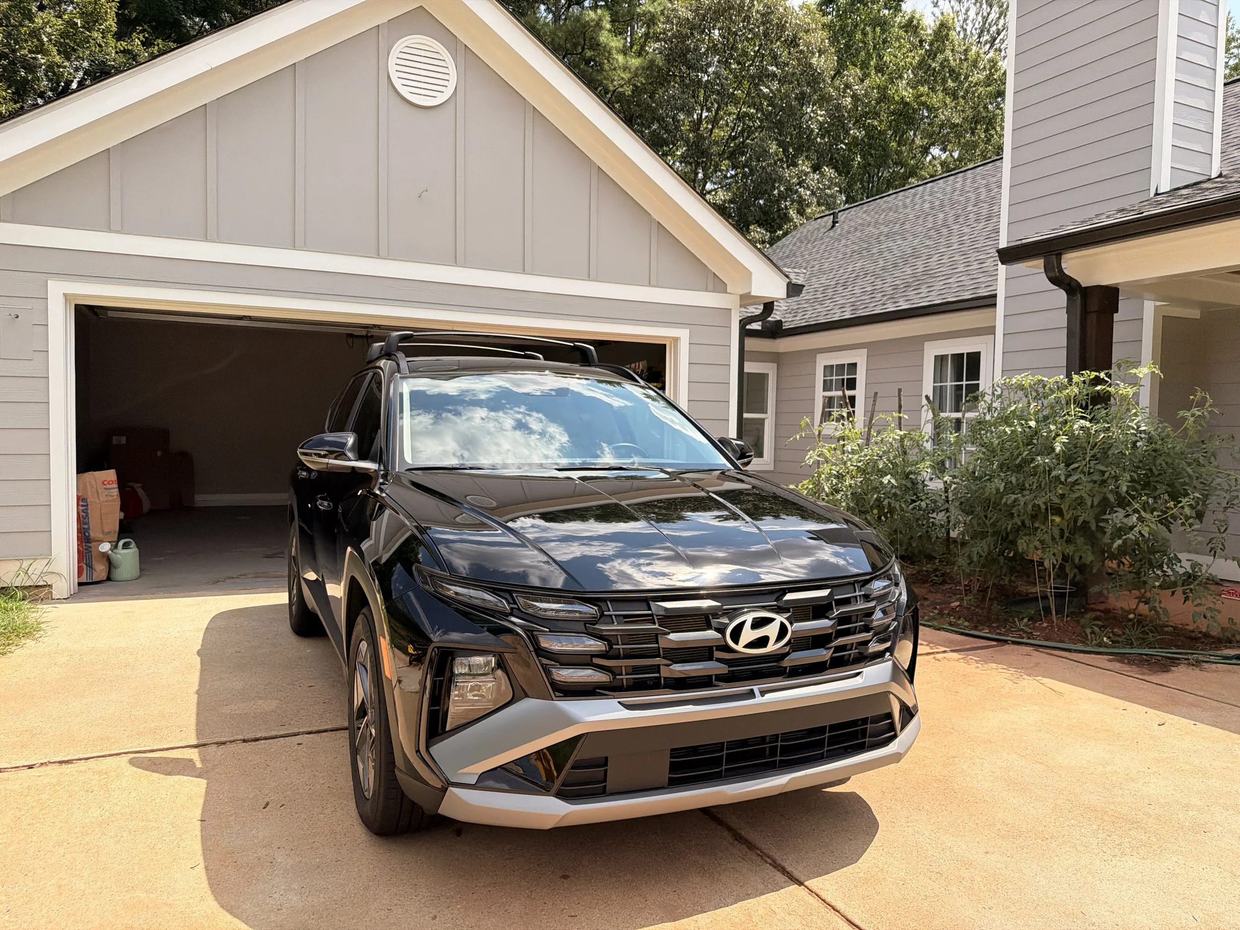 A black Hyundai SUV parked in the driveway of a gray house with white trim. The house has a garage with an open door, some boxes and a watering can visible inside, and gray siding with a round vent near the roof peak. There are trees in the backgroun