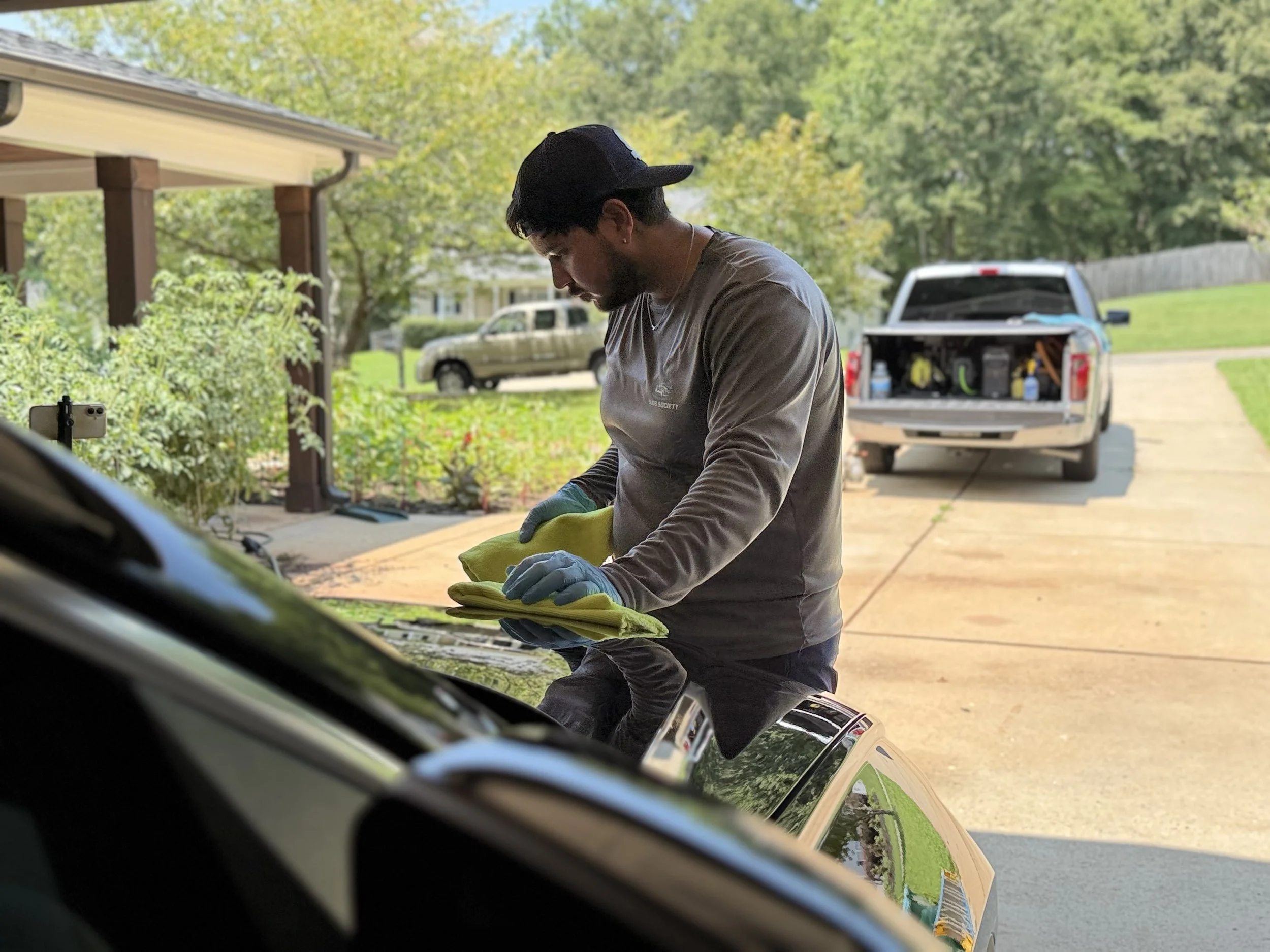 A man wearing a black cap and a gray long-sleeve shirt, with gloves, is cleaning or working on a car in a driveway. In the background, a pickup truck with its tailgate open, tools, and bottles are visible. The scene is outdoors with green trees and a