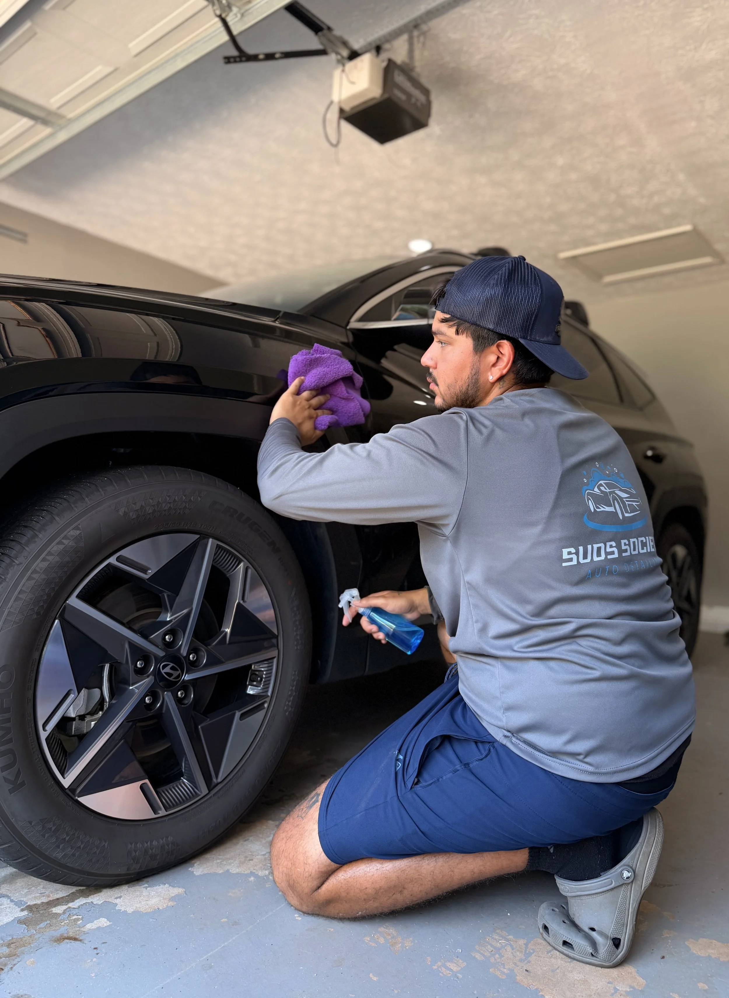 Man kneeling beside a black Hyundai vehicle, cleaning or polishing the tire with a purple cloth, holding a spray bottle, in a garage.
