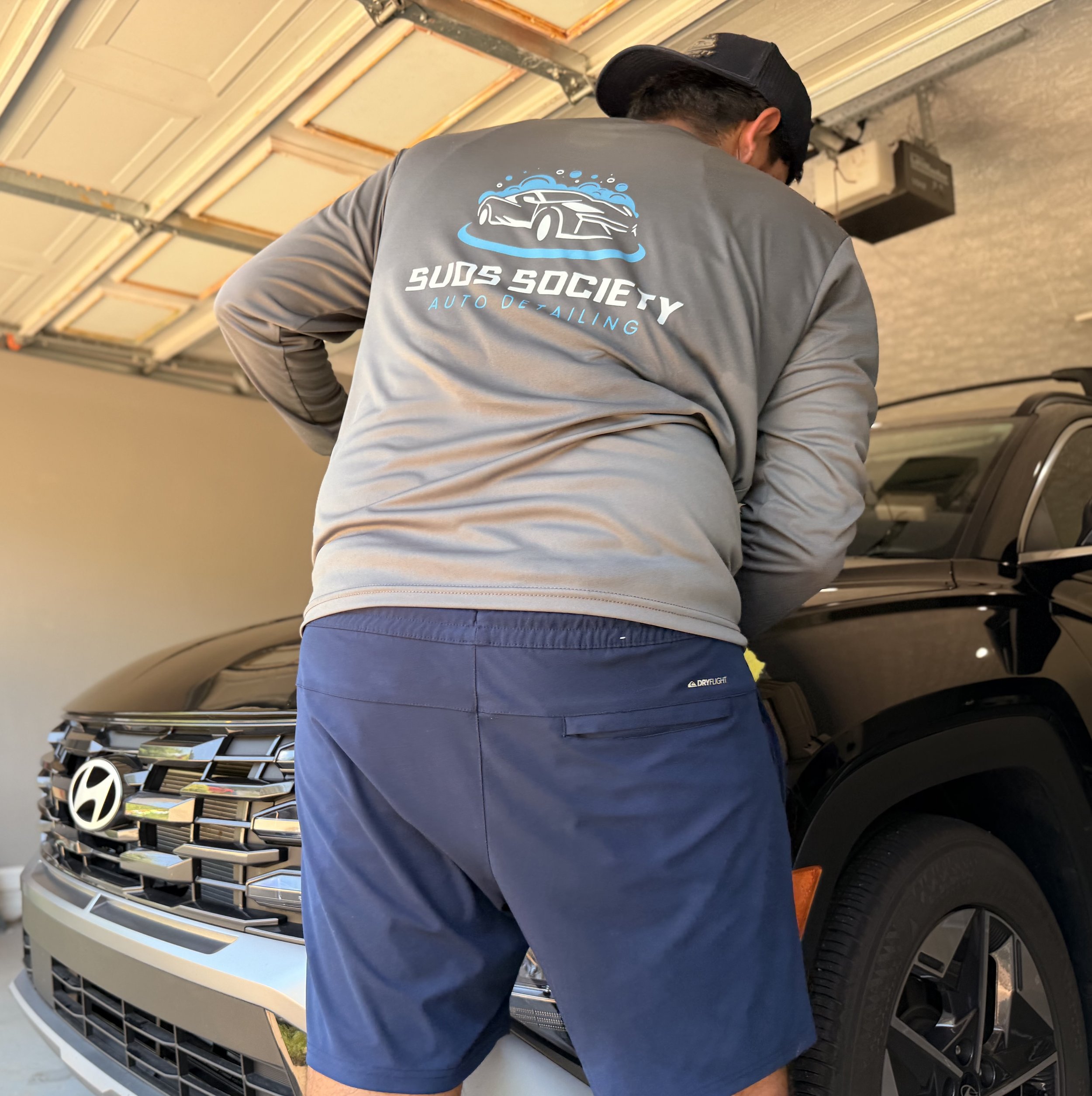 A man wearing a gray jacket with a logo and the text 'Suds Society Auto Detailing' and a black baseball cap is cleaning or inspecting the front grille and bumper of a gray SUV in a garage.