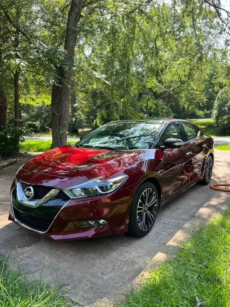 A red Nissan sedan parked on a concrete driveway surrounded by green grass and trees.
