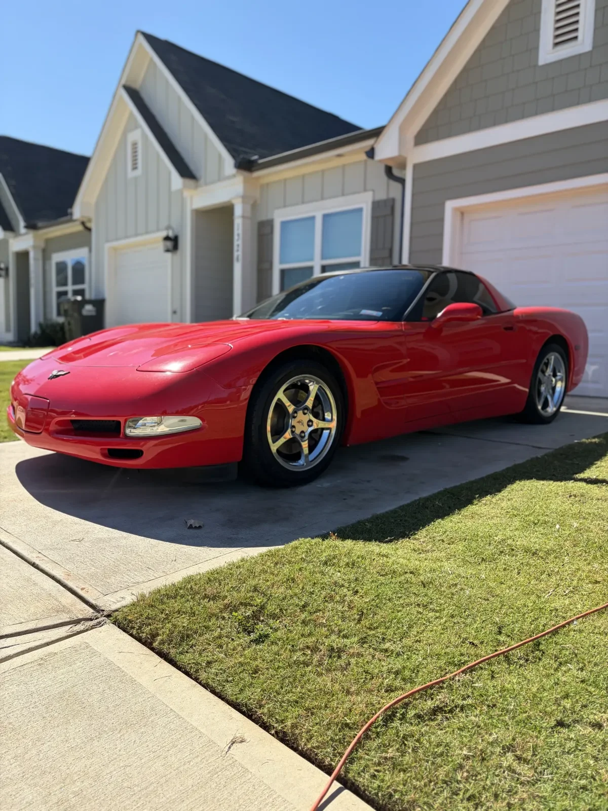 Red Chevrolet Corvette parked in front of suburban houses with garages, on a concrete driveway and lawn, under clear blue sky.