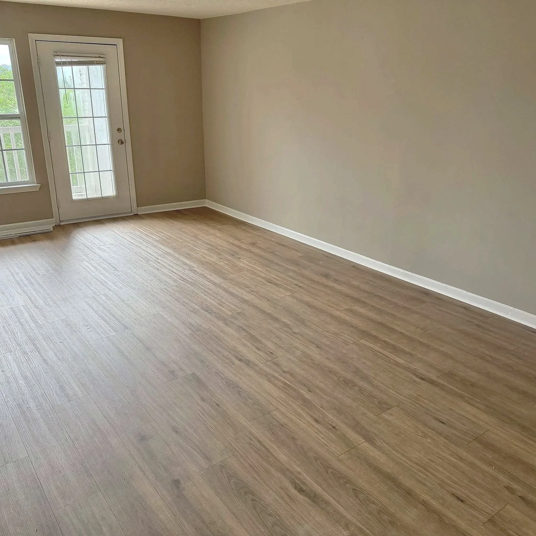Empty room with beige walls, wooden flooring, a glass door leading outside, and a single window with blinds.
