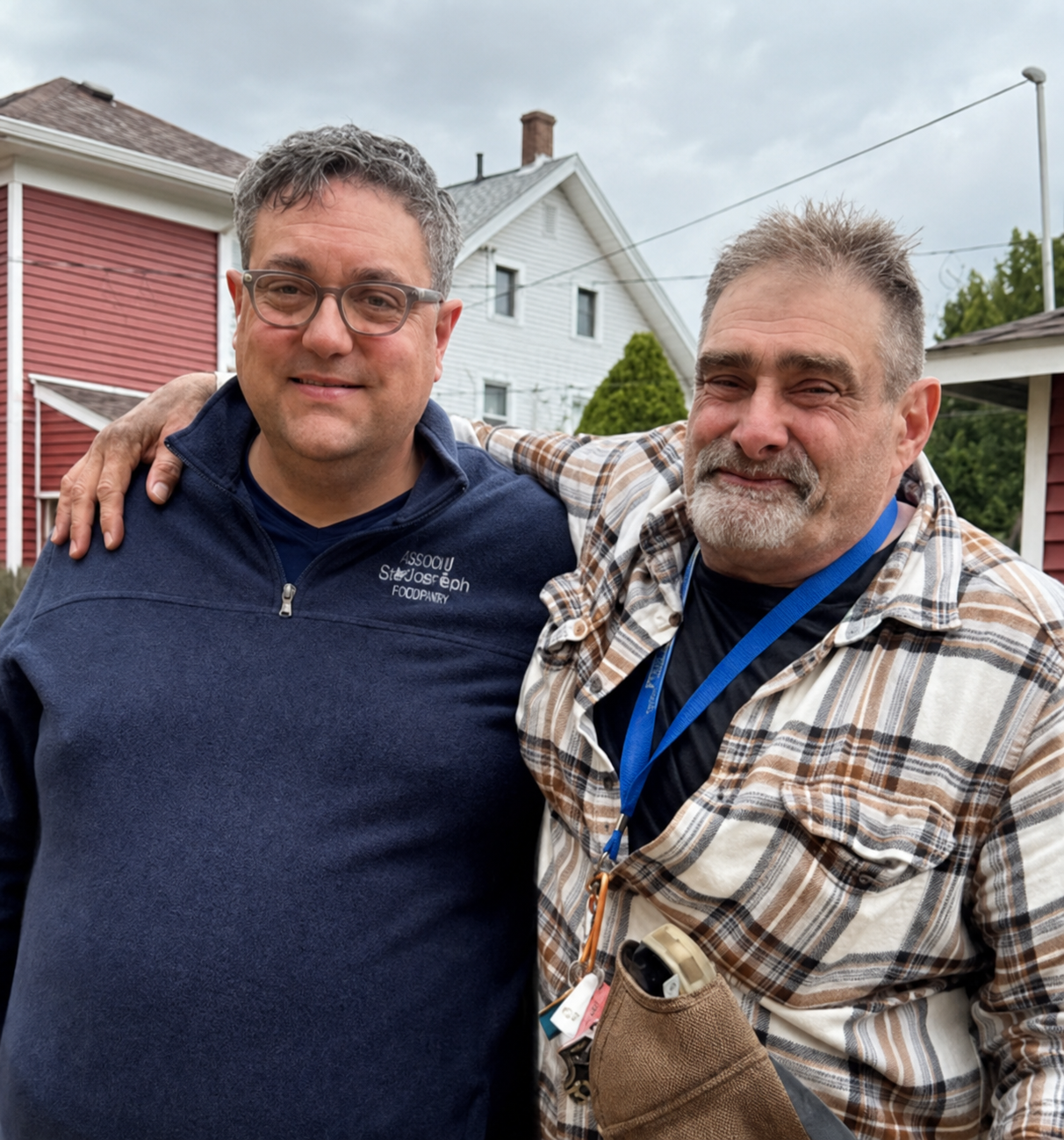 Two middle-aged men standing outdoors, smiling and with arms around each other's shoulders. One man wears glasses, a blue zip-up jacket, and the other wears a plaid shirt and has a beard. They are in front of houses and trees on a cloudy day.