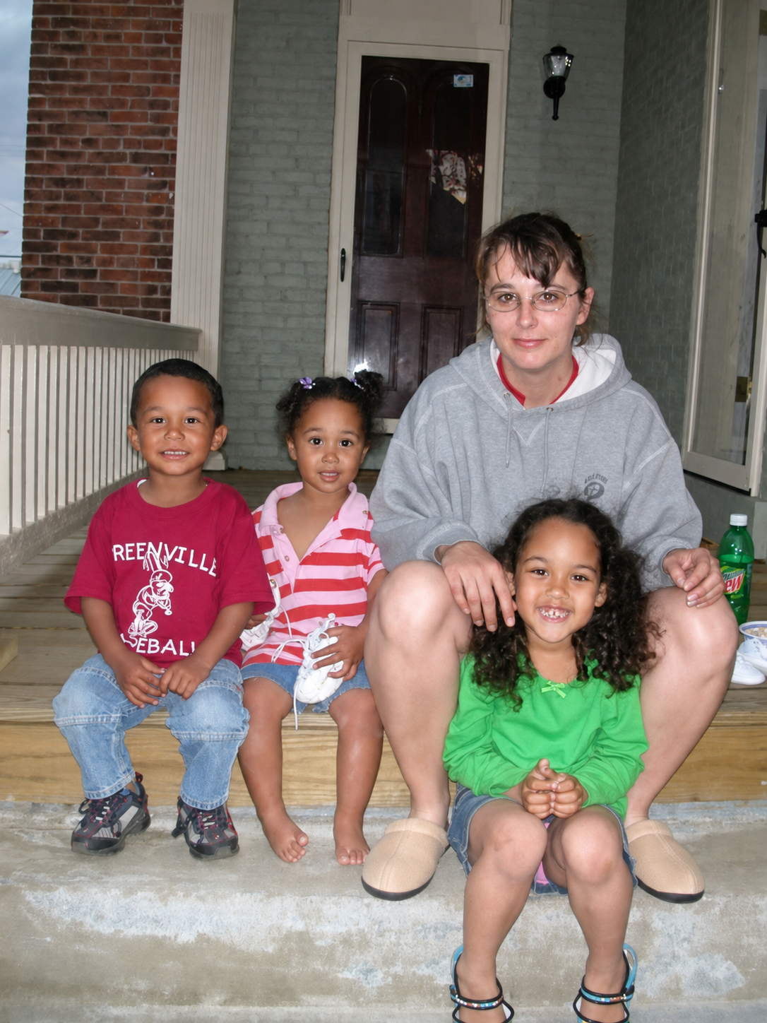 Four children and one woman sitting on a porch step. The woman is in a gray hoodie, and the children are in colorful casual clothing. They are smiling and sitting outdoors in front of a house.