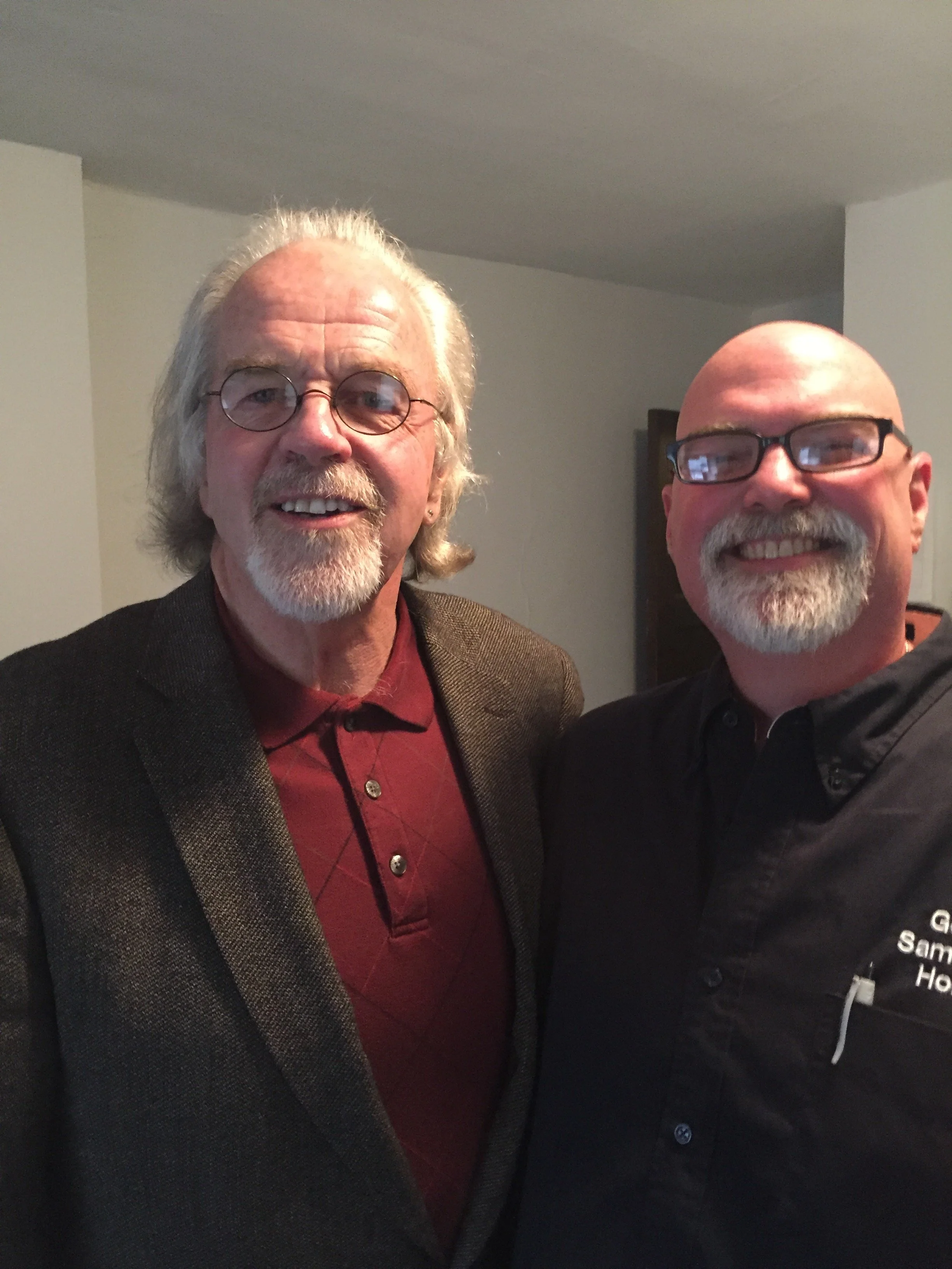 Two smiling men with glasses, our founder John David Graham and executive director Brian Gilvin, one with white hair and beard, and the other with a bald head and mustache, taking a selfie together indoors.