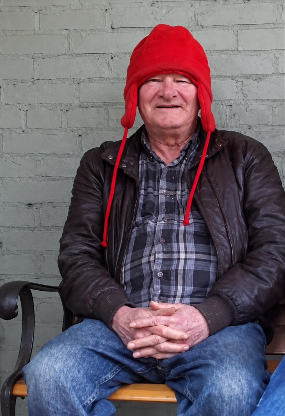 An elderly man sitting on a bench wearing a red winter hat with ear flaps, a leather jacket, a plaid shirt, and jeans against a gray brick wall.