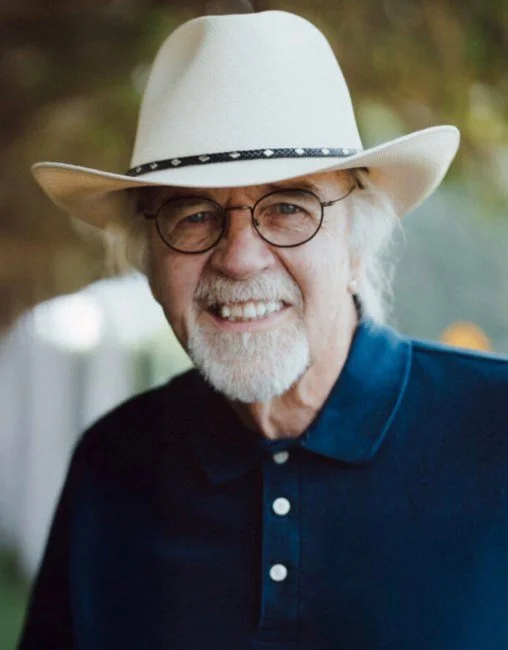 John David Graham, the Good Samaritan Home founder, with glasses and a beard wearing a white cowboy hat and a dark blue collared shirt, smiling outdoors.