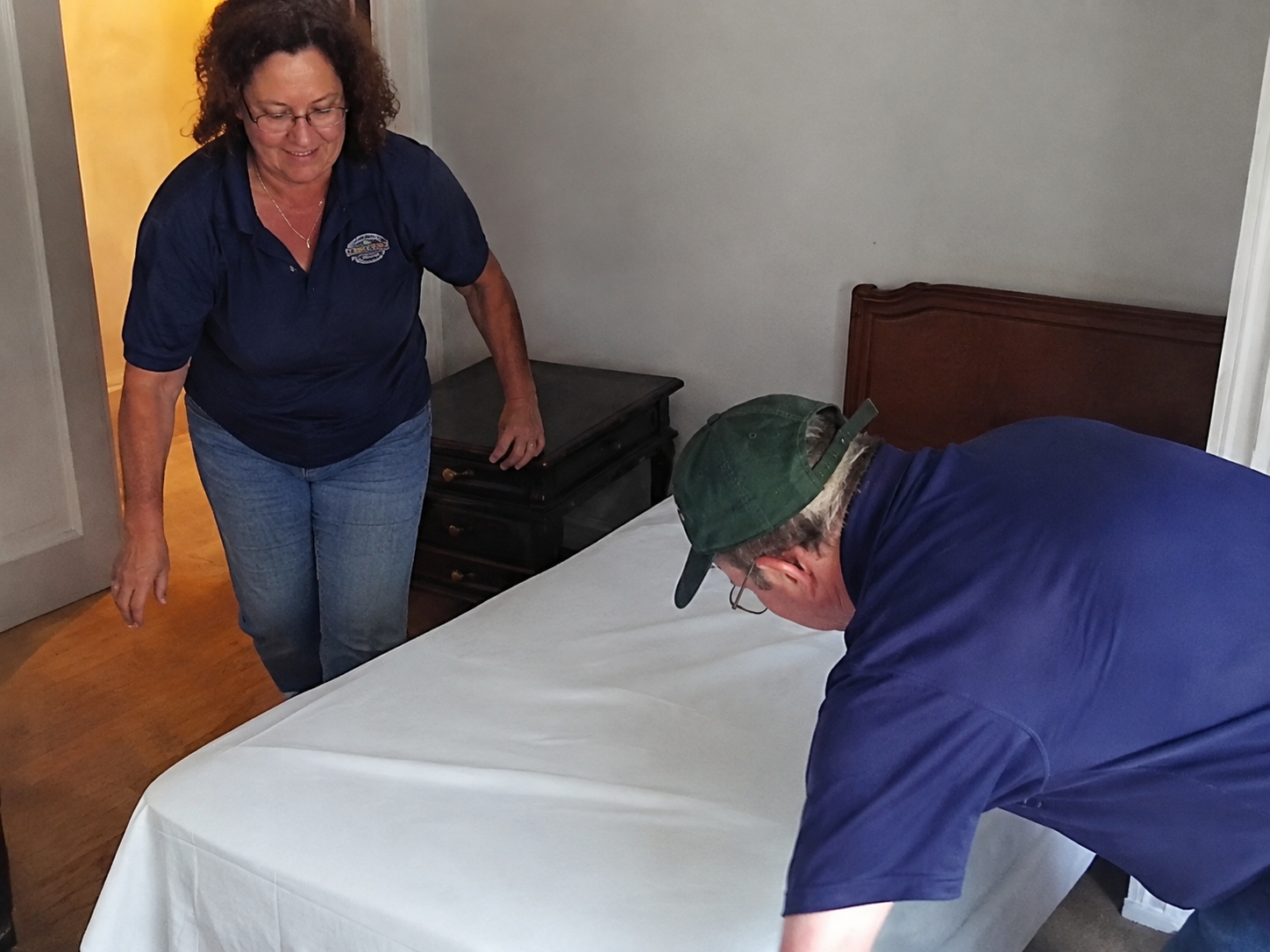 A woman and a man are making the bed in a bedroom. The woman is standing and looking down at the bed, while the man is bent over and smoothing a white bedsheet. The woman has curly hair and is wearing glasses, a navy blue polo shirt, and blue jeans. The man is wearing a green backwards cap, glasses, and a navy blue shirt.