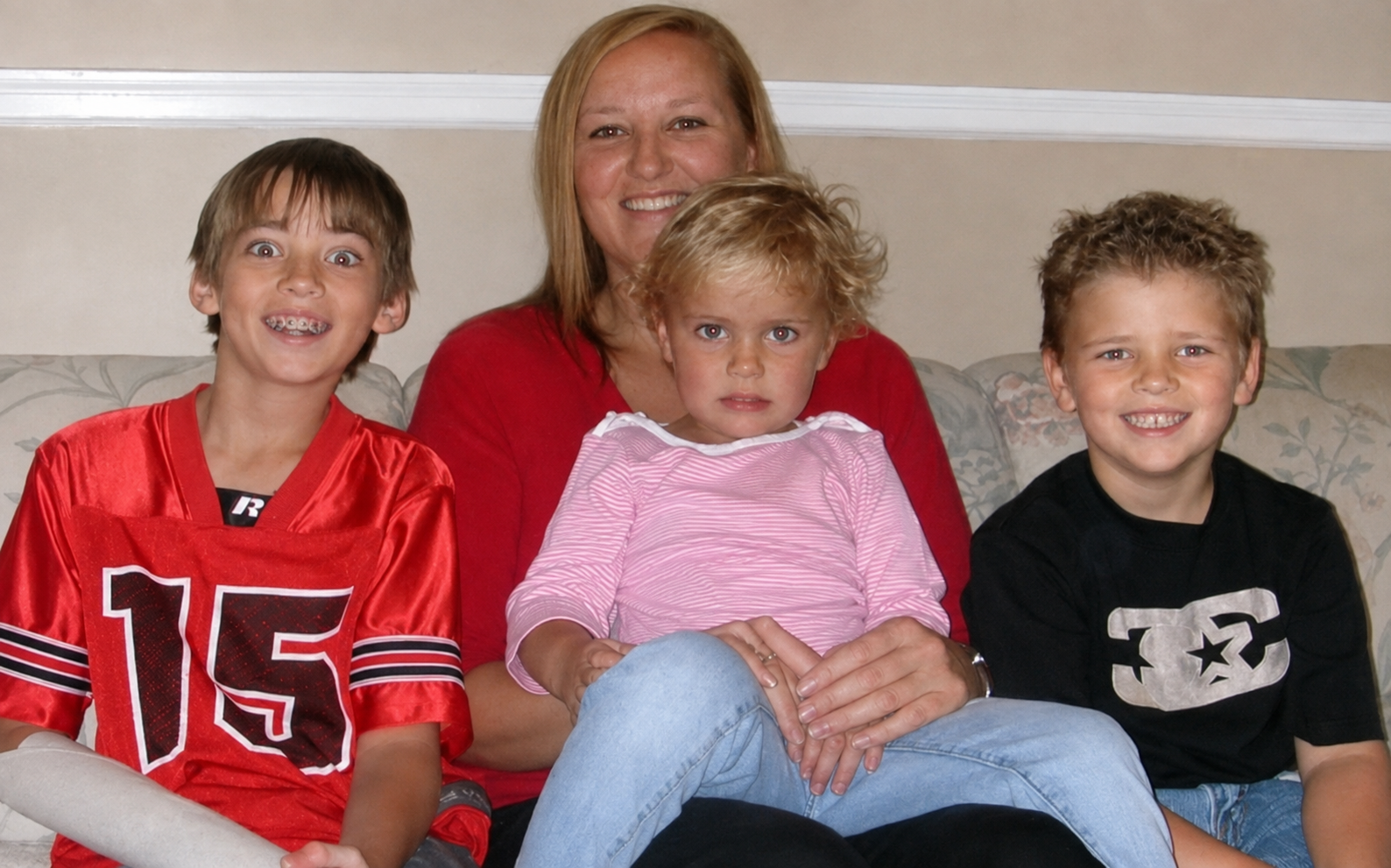 A smiling woman sitting on a floral sofa with three young children, two boys and a girl, in a living room. The girl, sitting in the middle, has curly blonde hair and is wearing a pink and white striped long sleeve shirt. The boy on the left has straight light brown hair, braces, and is wearing a red sports jersey with the number 15. The boy on the right has curly blonde hair and is wearing a black shirt with a silver design.