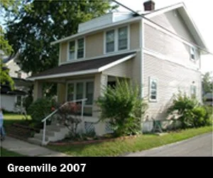 A two-story beige house with a front porch and stairs, green bushes and trees in front, and a sidewalk in a residential neighborhood depicting Good Samaritan Home's housing expansion for those looking for re-entry. 