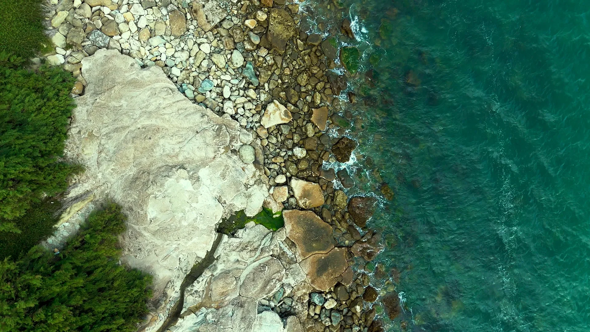 Aerial view of a rocky shoreline with a concrete cliff on the left and green trees on the far left, meeting the turquoise ocean with waves washing over rocks.