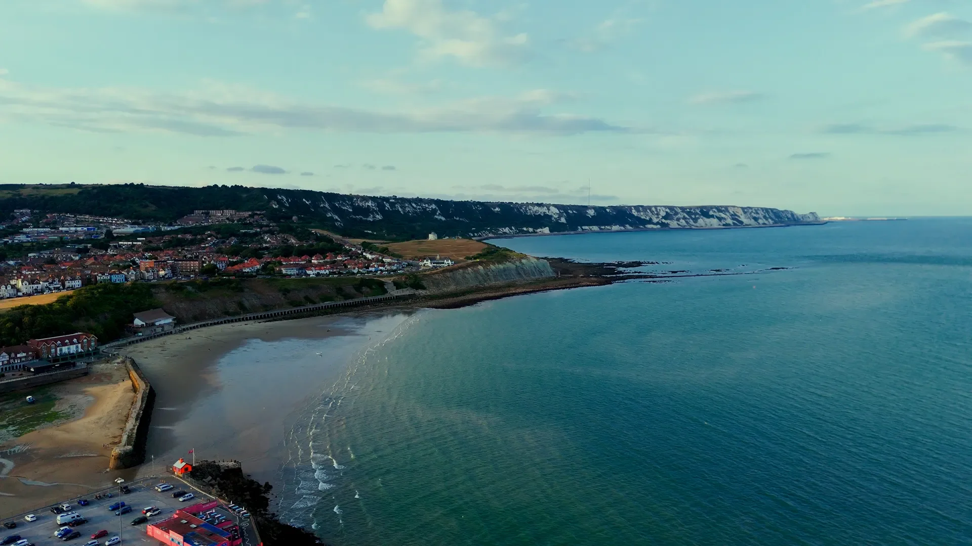 A coastal scene with houses on cliffs, a beach with a pier, and the ocean with gentle waves, under a partly cloudy sky.