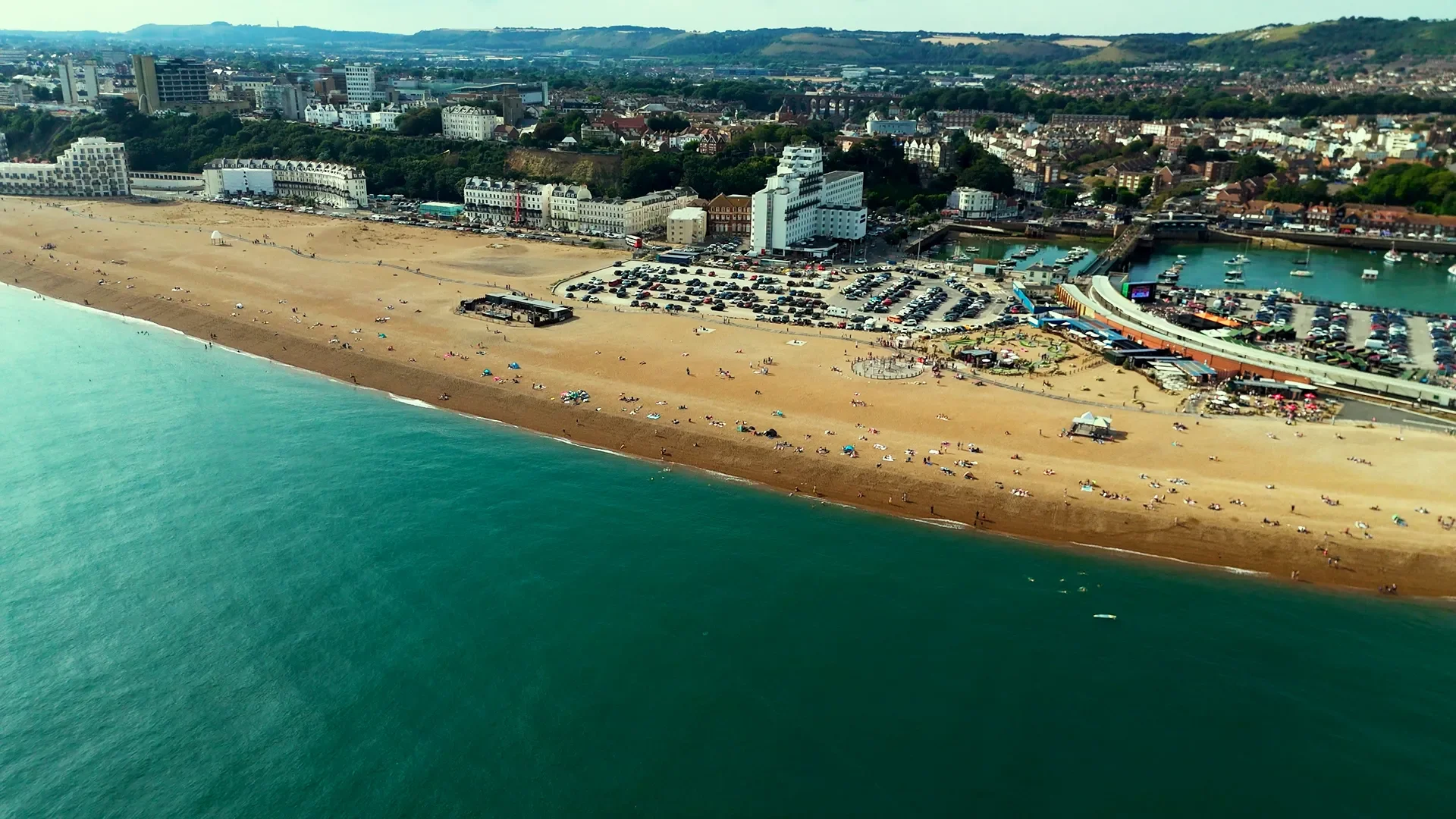 Aerial view of a beach with people sunbathing and walking, adjacent to a parking lot and buildings near a marina, with hilly terrain in the background.