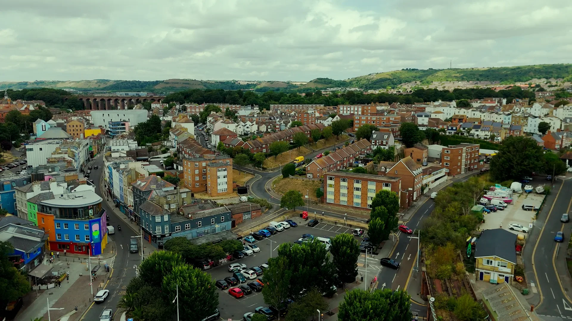 A cityscape view with a mix of colorful buildings, green trees, cars parked along streets, and hills in the background under partly cloudy skies.