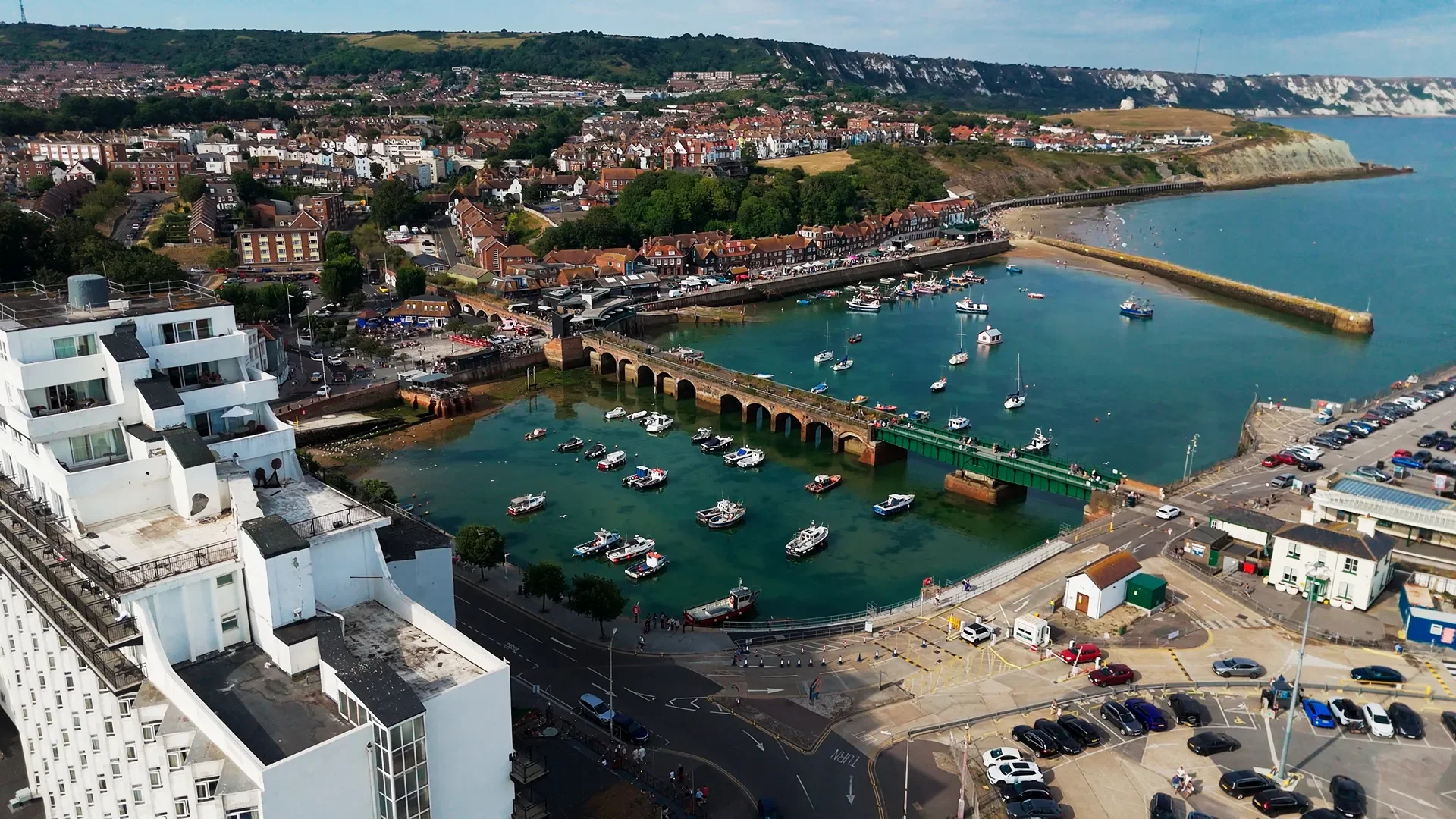 Aerial view of a coastal town with a harbor filled with boats and yachts. There are residential and commercial buildings, with a harbor wall extending into the water. The background features green hills and white cliffs along the coast.