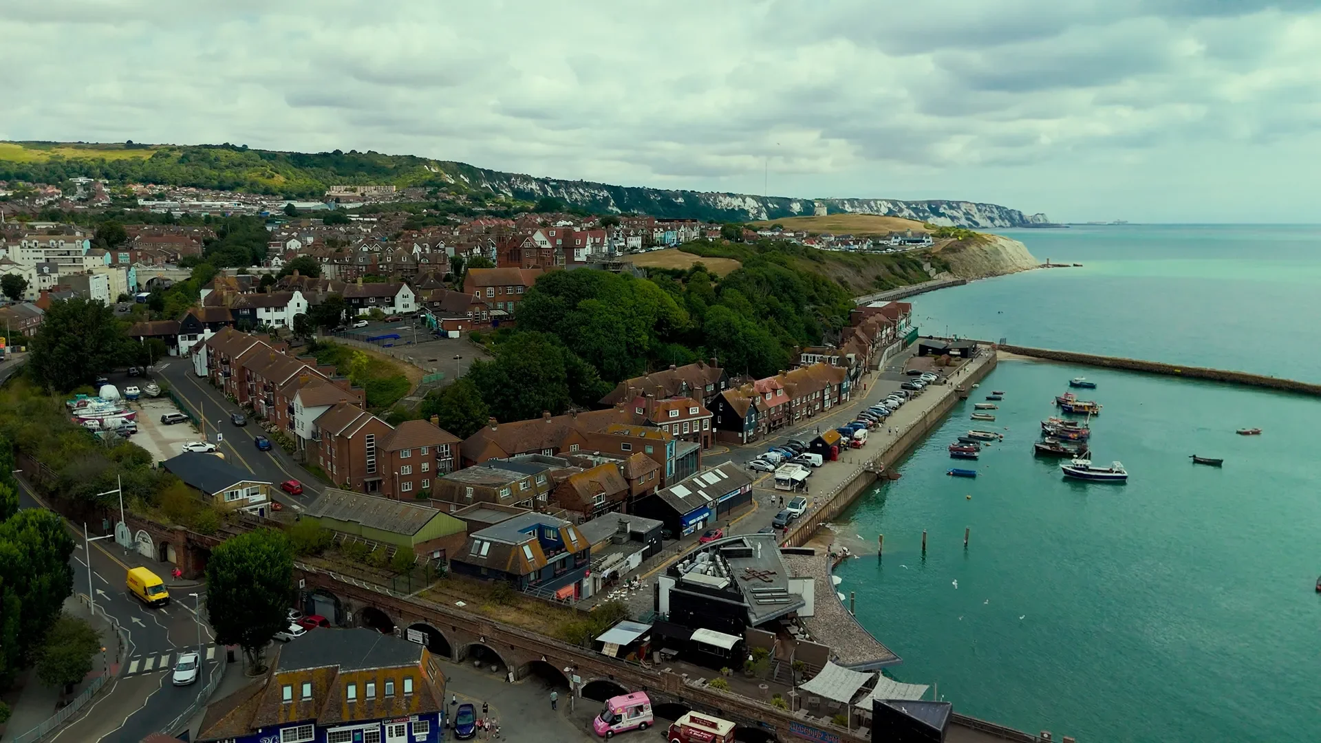 A coastal town with a harbor filled with boats, surrounding buildings, and street traffic, with green hills and cliffs in the distance under a cloudy sky.