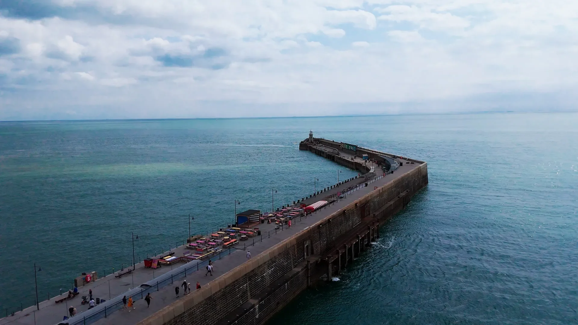 A long pier extends into the ocean with a small lighthouse at the end. There are people walking along the pier and sitting at tables near the entrance. The sky is partly cloudy, and the water is calm.