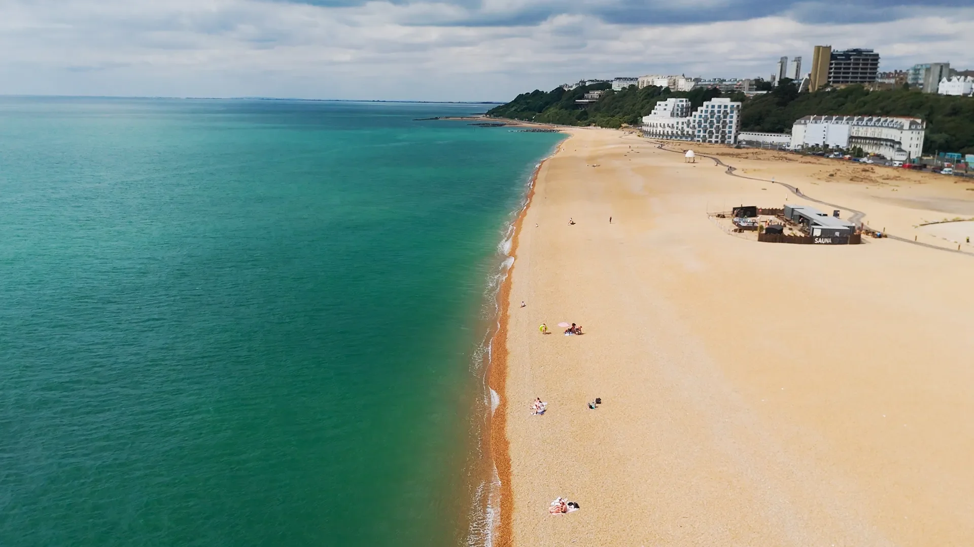 Aerial view of a beach with turquoise water on the left and sandy shore on the right, with a few people relaxing under umbrellas and some walking along the beach. Buildings and greenery are visible in the background.
