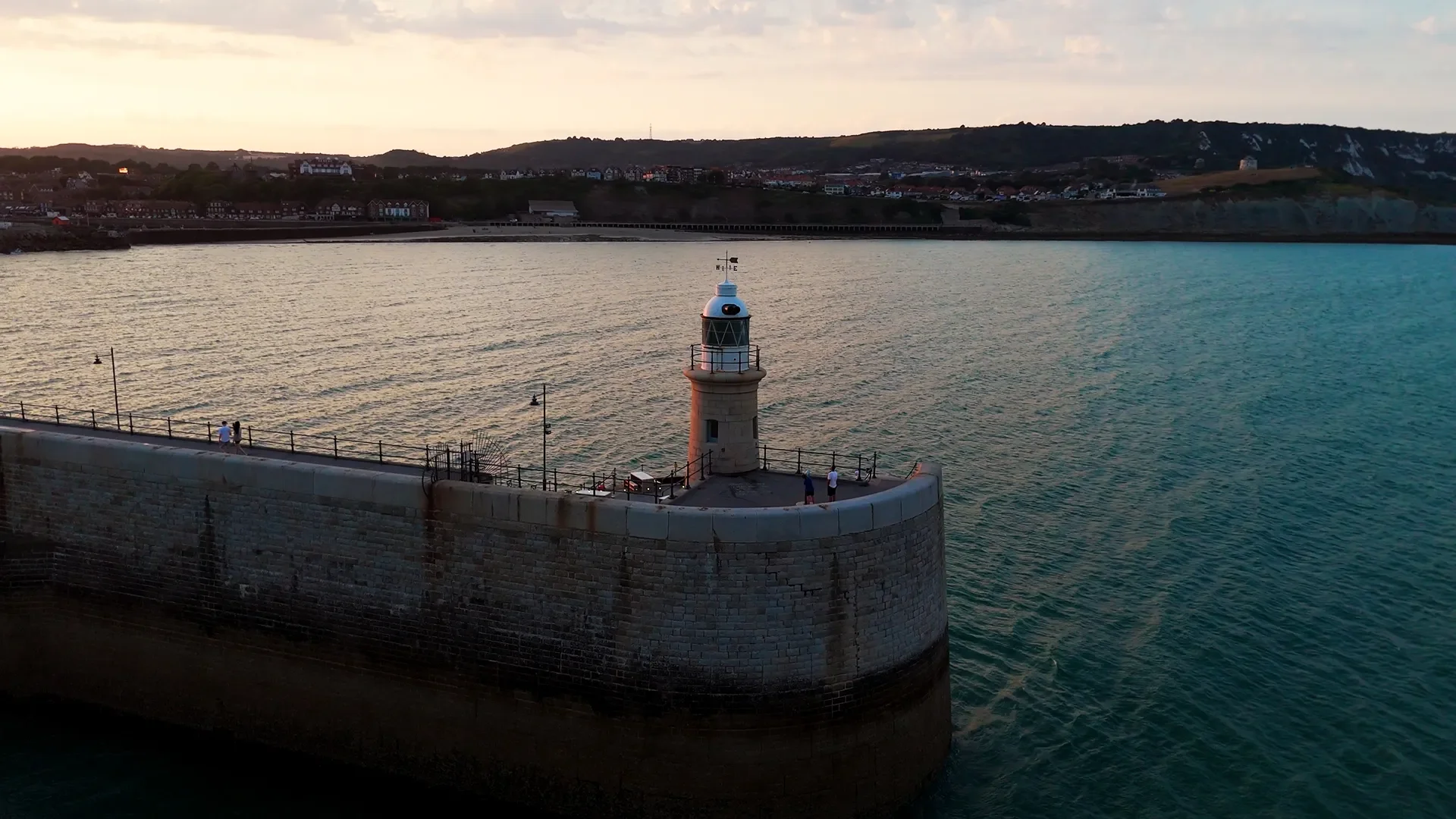 A harbor with a stone pier and a lighthouse at the end, overlooking the water and distant hills with buildings and houses, under a partly cloudy sky.