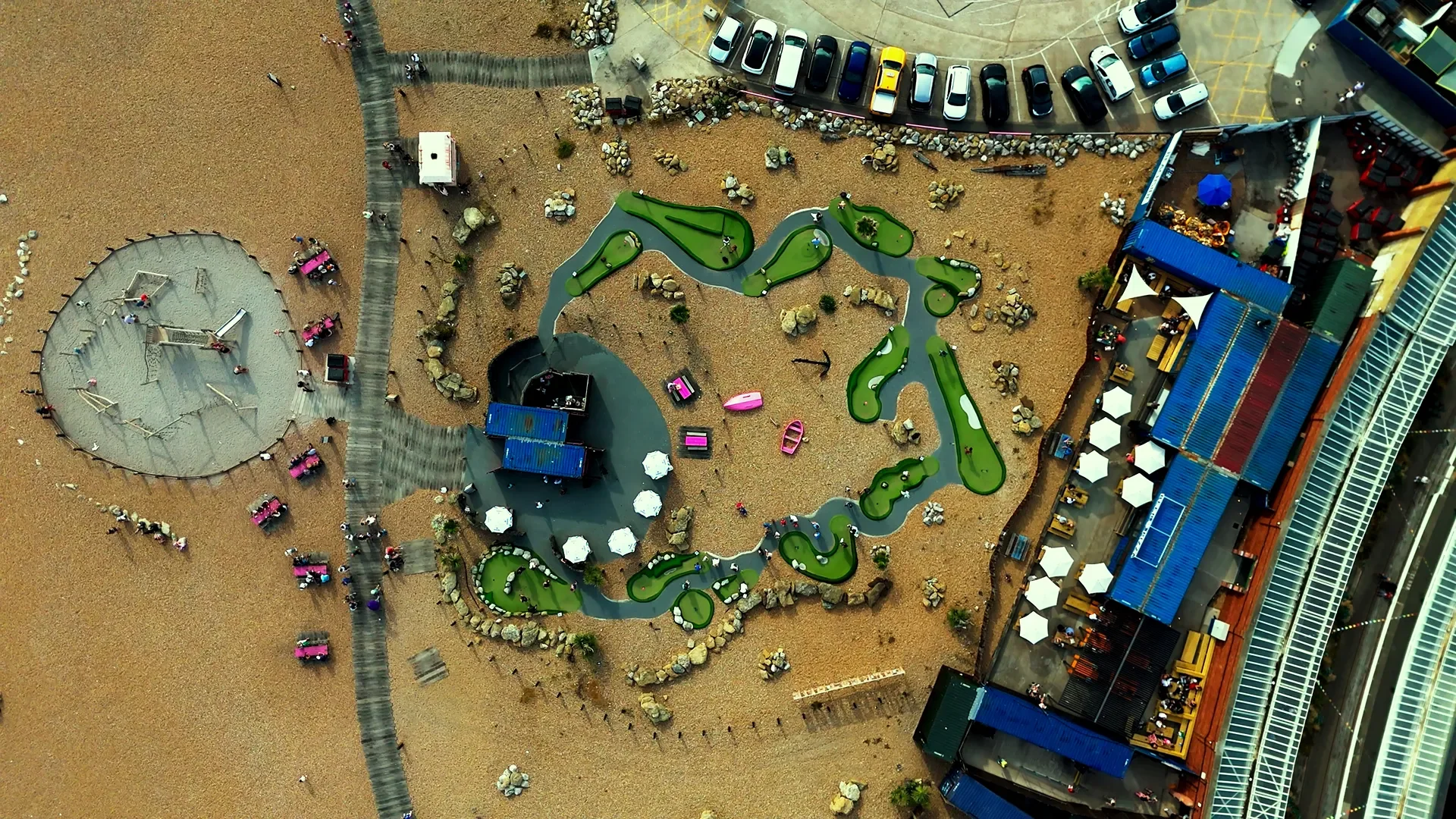 An aerial view of a mini golf course, parking lot, and surrounding areas with people walking and colorful umbrellas.