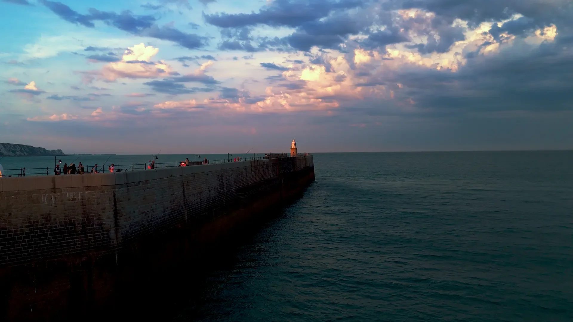 Seaside pier with people fishing and walking, a lighthouse at the end, and a dramatic sky with clouds at sunset.