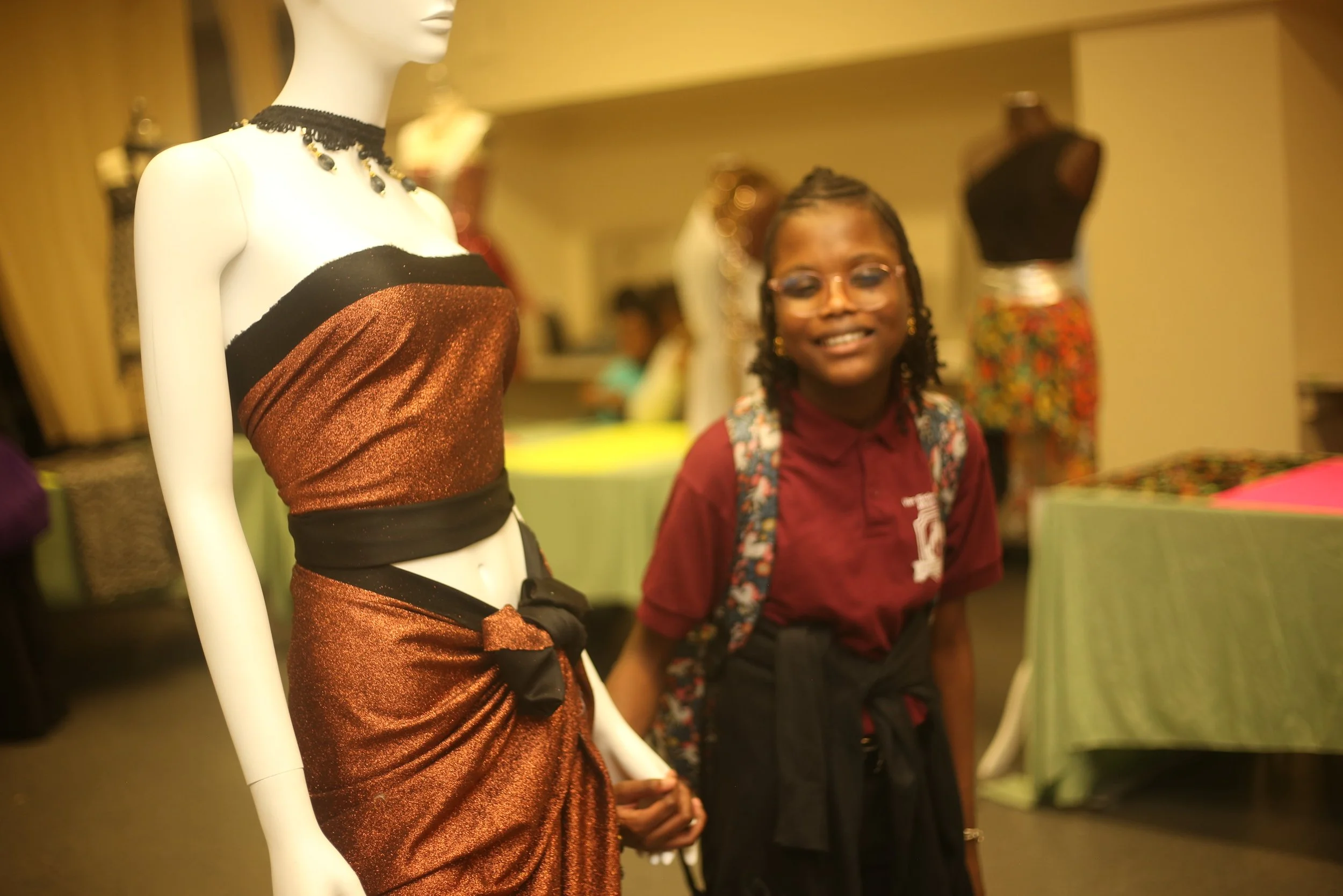 A mannequin dressed in a metallic, copper-colored tube top and matching skirt with black trim, standing next to a smiling young woman with glasses, braided hair, and a maroon shirt with a patterned backpack, in an indoor retail space with display man
