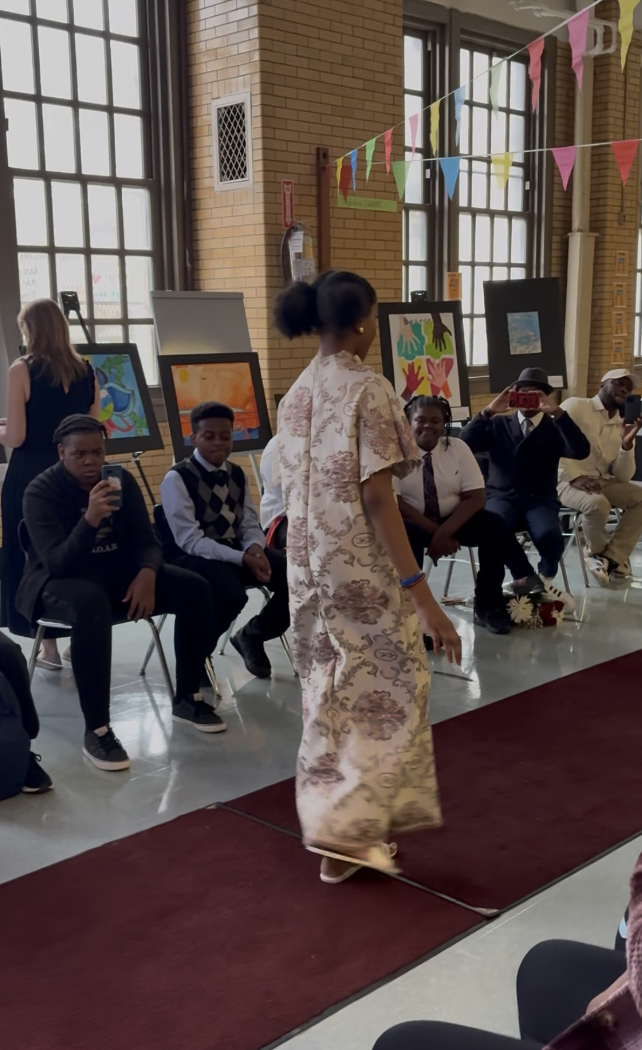 A woman in traditional dress walking on a red carpet in a room with brick walls and large windows. Behind her, several people are seated, some taking photos or videos. There are colorful banners hanging from the ceiling and framed artwork displayed o