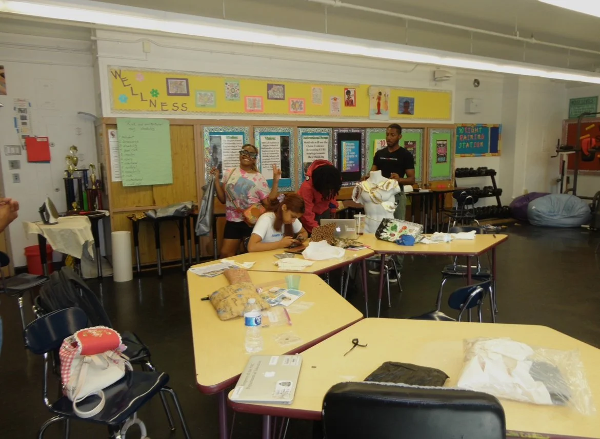 A group of women and a man working on a fashion project in a classroom with tables, a mannequin, and various craft supplies and decorations.