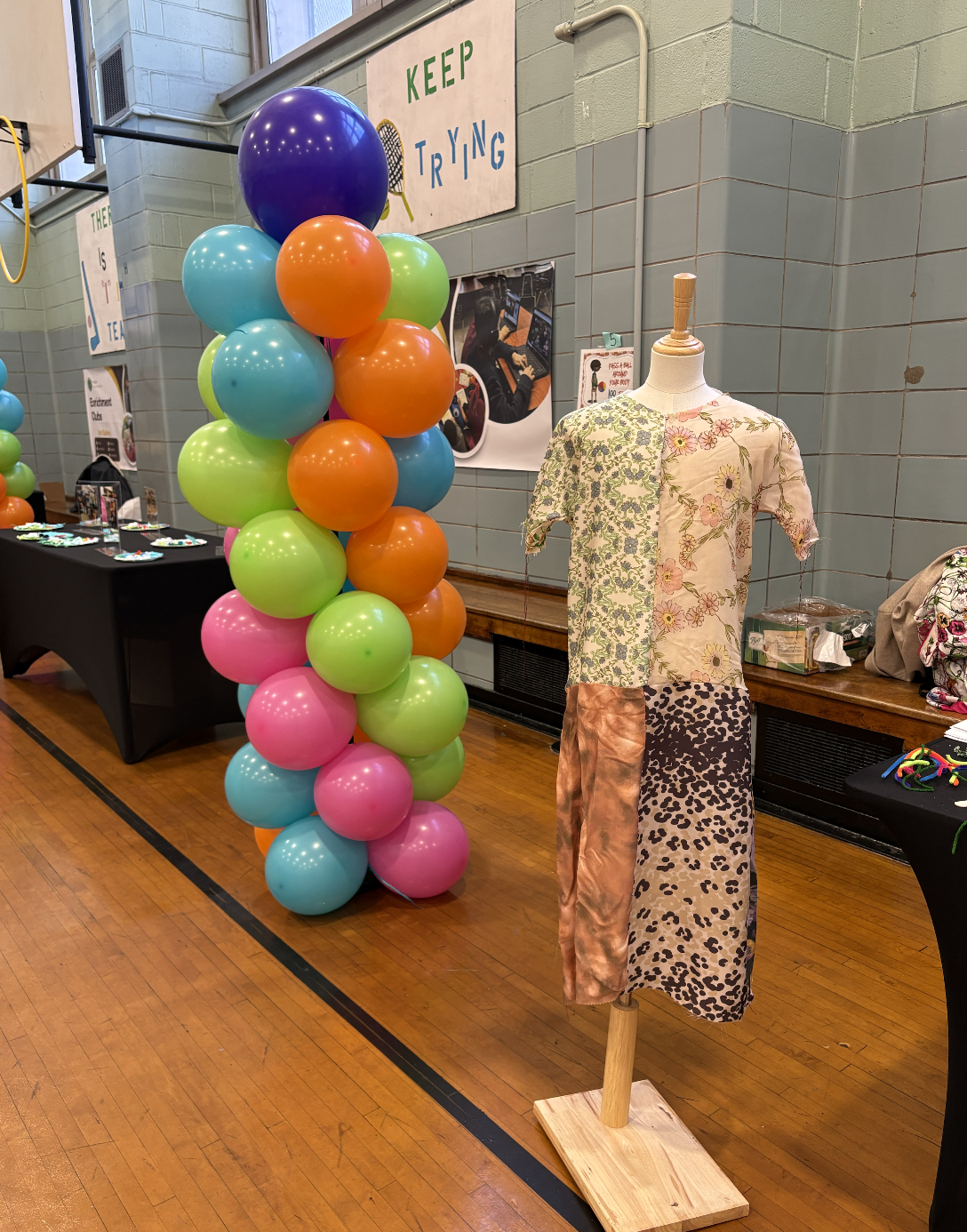 Colorful balloons arranged in a column standing next to a mannequin dressed in a patchwork-style dress with floral and animal print patterns. The scene appears to be at an indoor event or fair with tables displaying items and signs in the background.