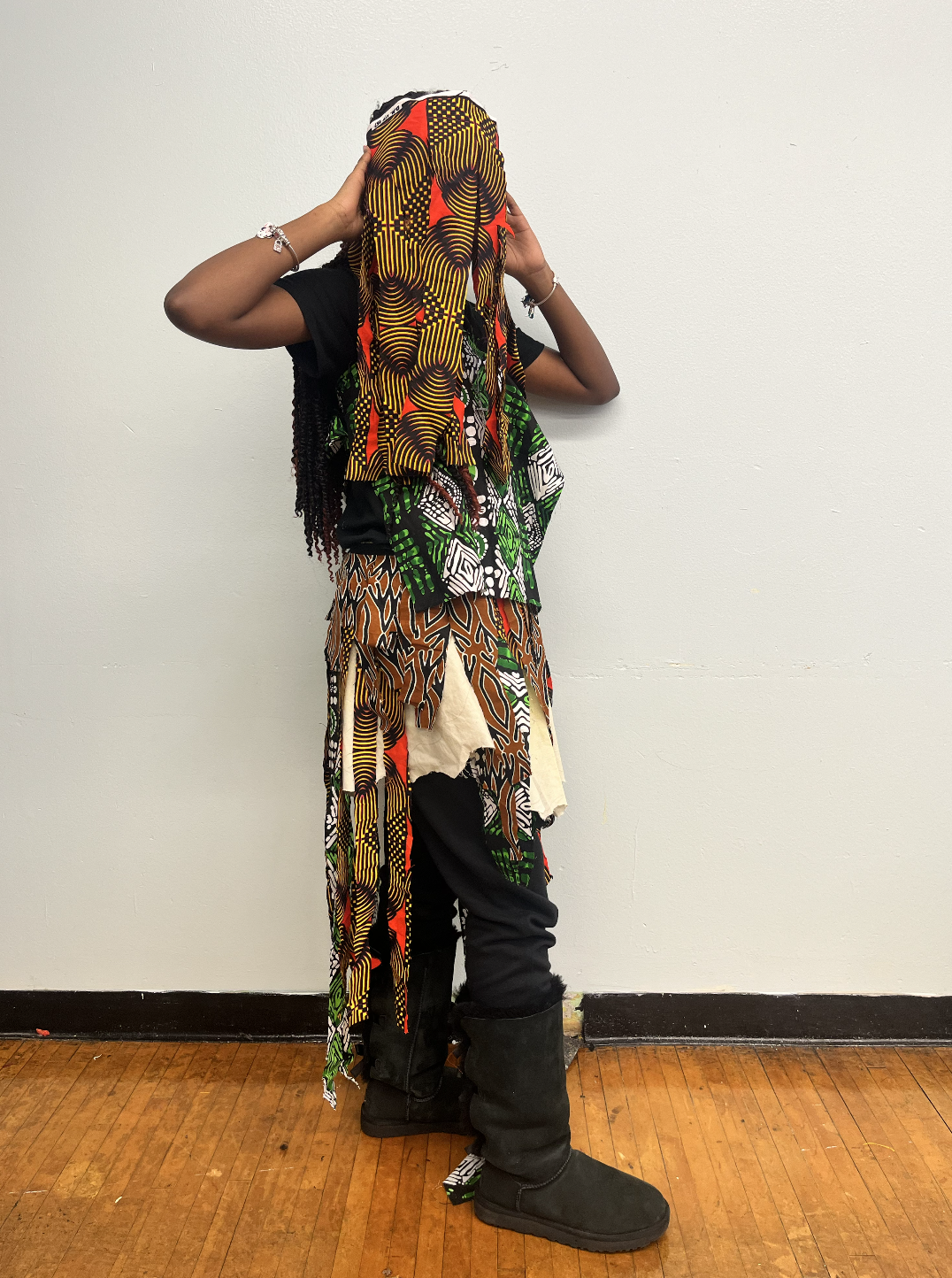 Person wearing a colorful African-patterned face mask and layered clothing standing against a plain white wall.