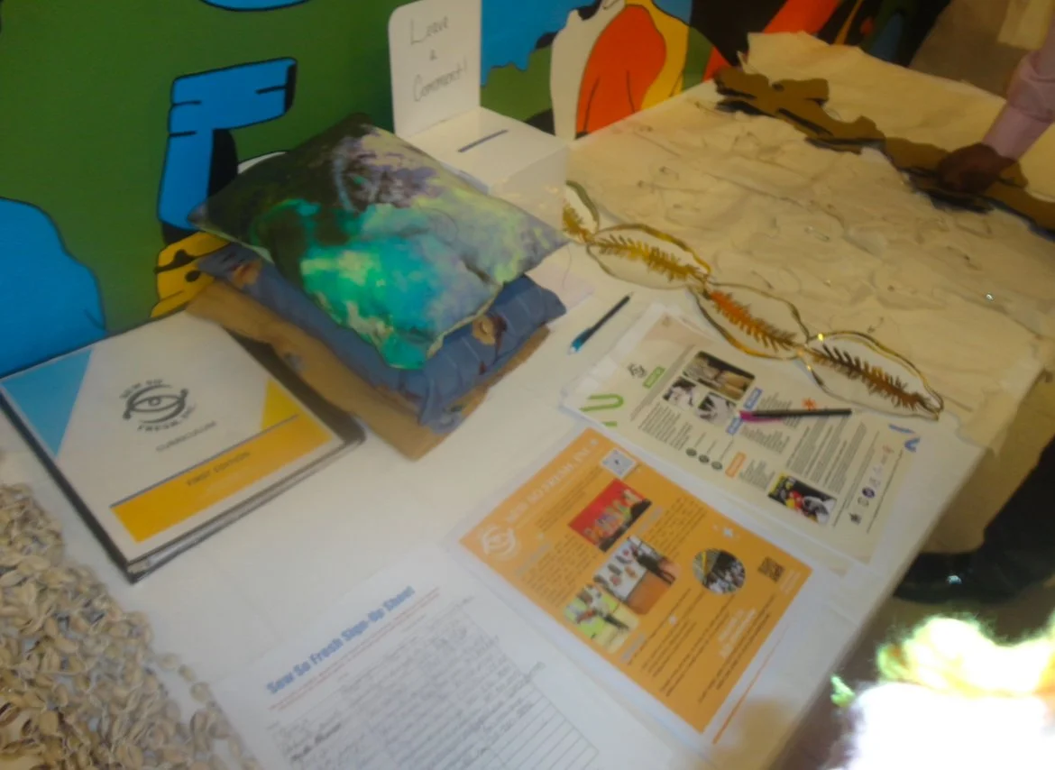 An informational display table with pamphlets, a pillow, and decorative leaves or vines, set against a colorful mural background.