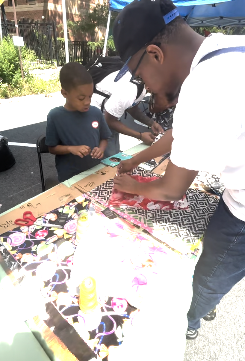 A man and a boy looking at fabric samples at an outdoor event table with colorful fabric swatches and sewing supplies.
