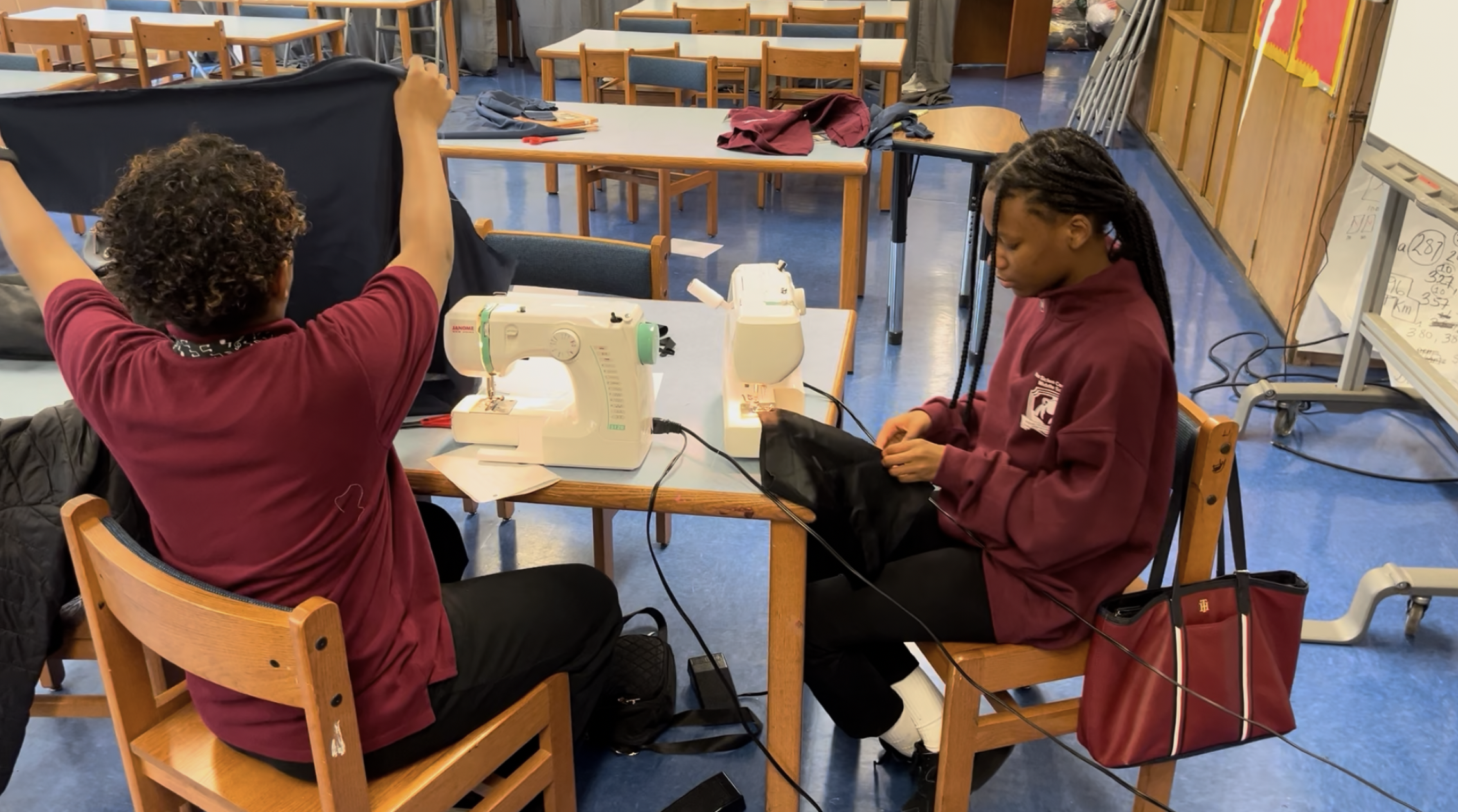Two students sewing at a table in a classroom, with sewing machines and fabric, surrounded by empty chairs and tables.