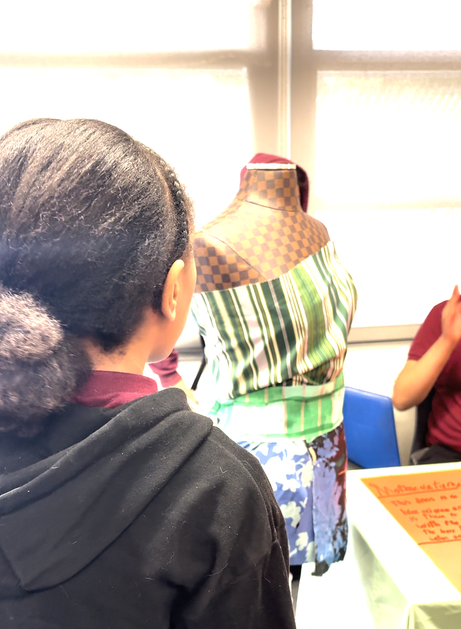 A person with dark hair in a ponytail looks at a fabric-covered mannequin wearing a green, striped, off-the-shoulder top, with two other people nearby, one sitting at a table.