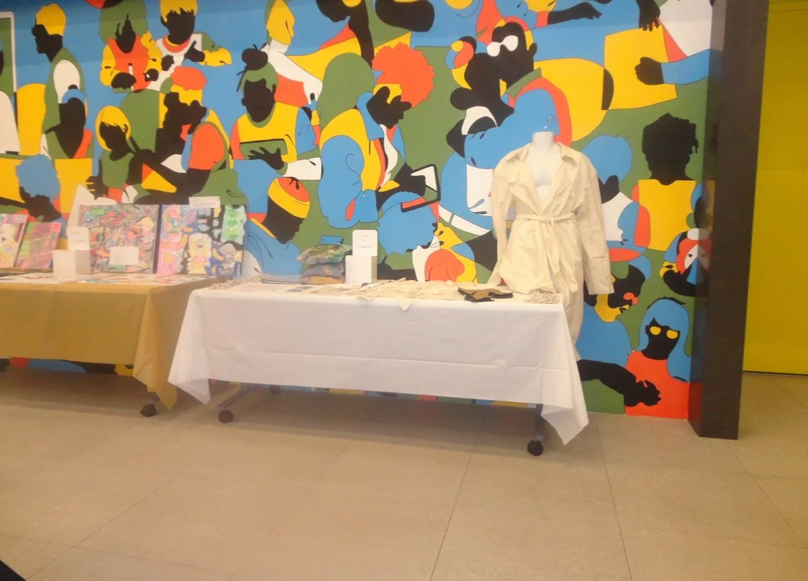 A display table with books and a mannequin dressed in a white garment, set against a colorful mural of abstract characters with diverse skin tones and hairstyles.