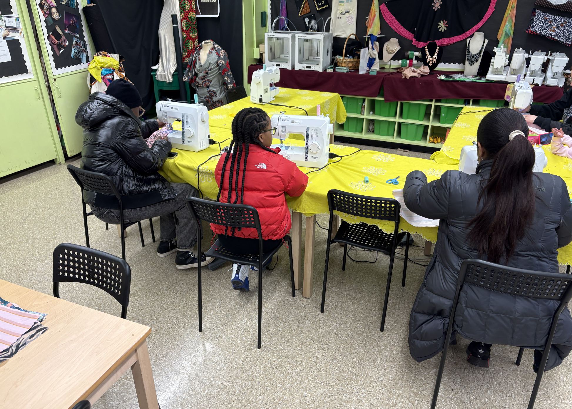 Children sitting at yellow-covered tables sewing on white sewing machines in a classroom or workshop, with sewing supplies, mannequins, photographs, and fabric displays in the background.