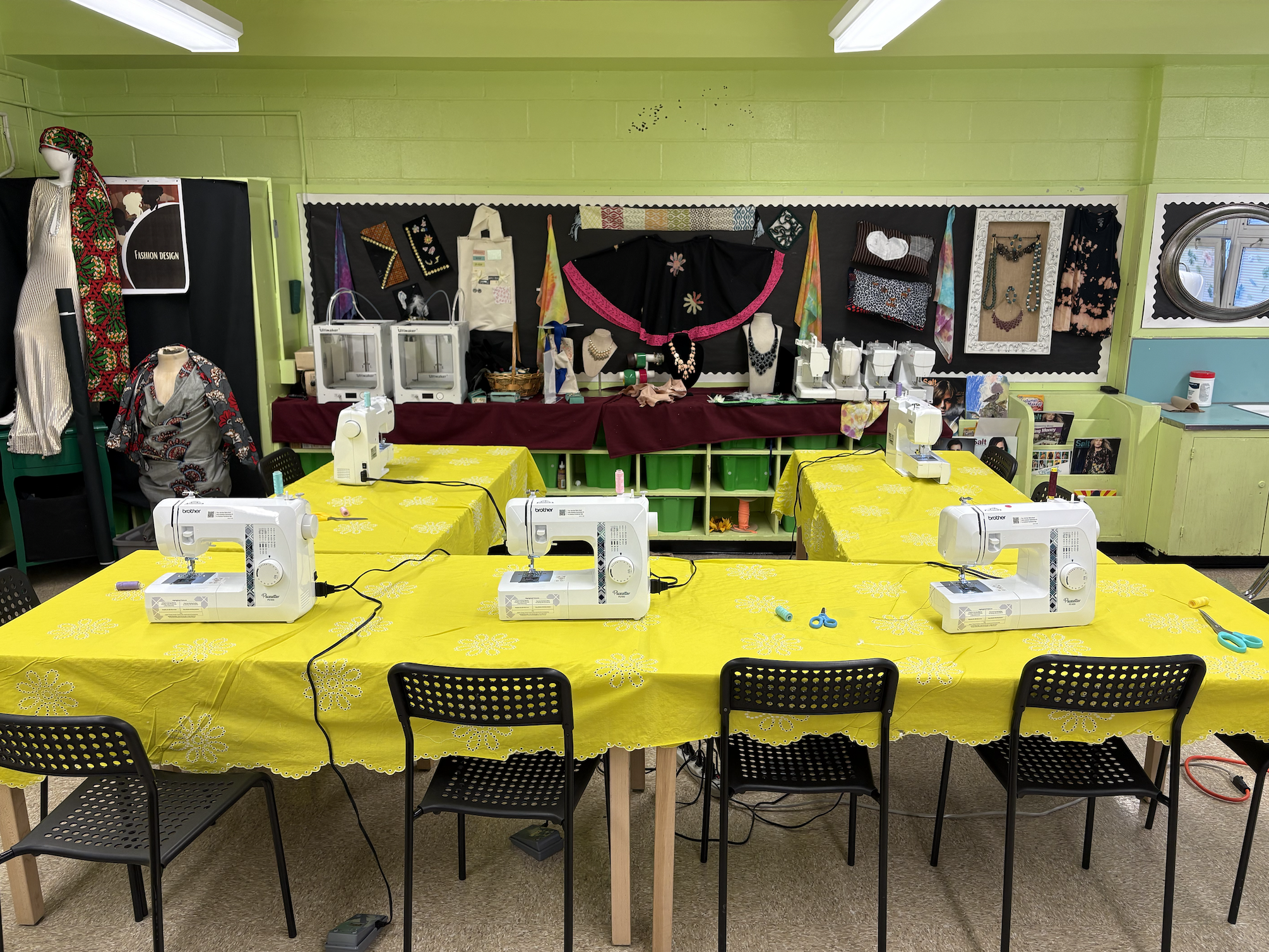 A classroom or craft room with several sewing machines on a yellow tablecloth-covered table, chairs around the table, and a wall decorated with fabric samples, jewelry, and other craft items, with sewing and craft supplies on a side table.