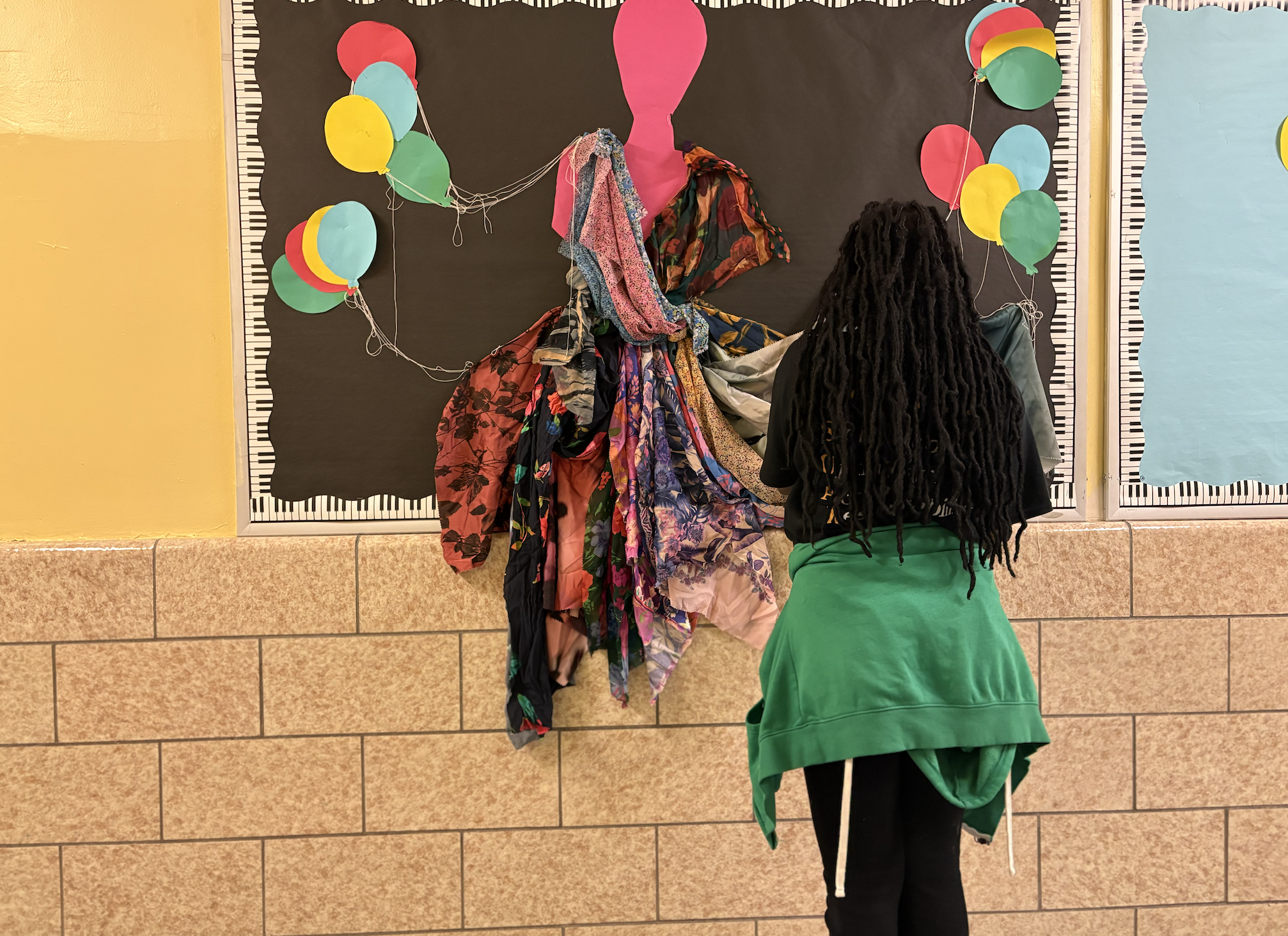 A person with long black dreadlocks wearing a green jacket and black pants is standing in front of a black bulletin board decorated with colorful balloons and a pink dress silhouette. The balloons are attached in a string, and various pieces of fabric are hanging on the board, resembling a dress.