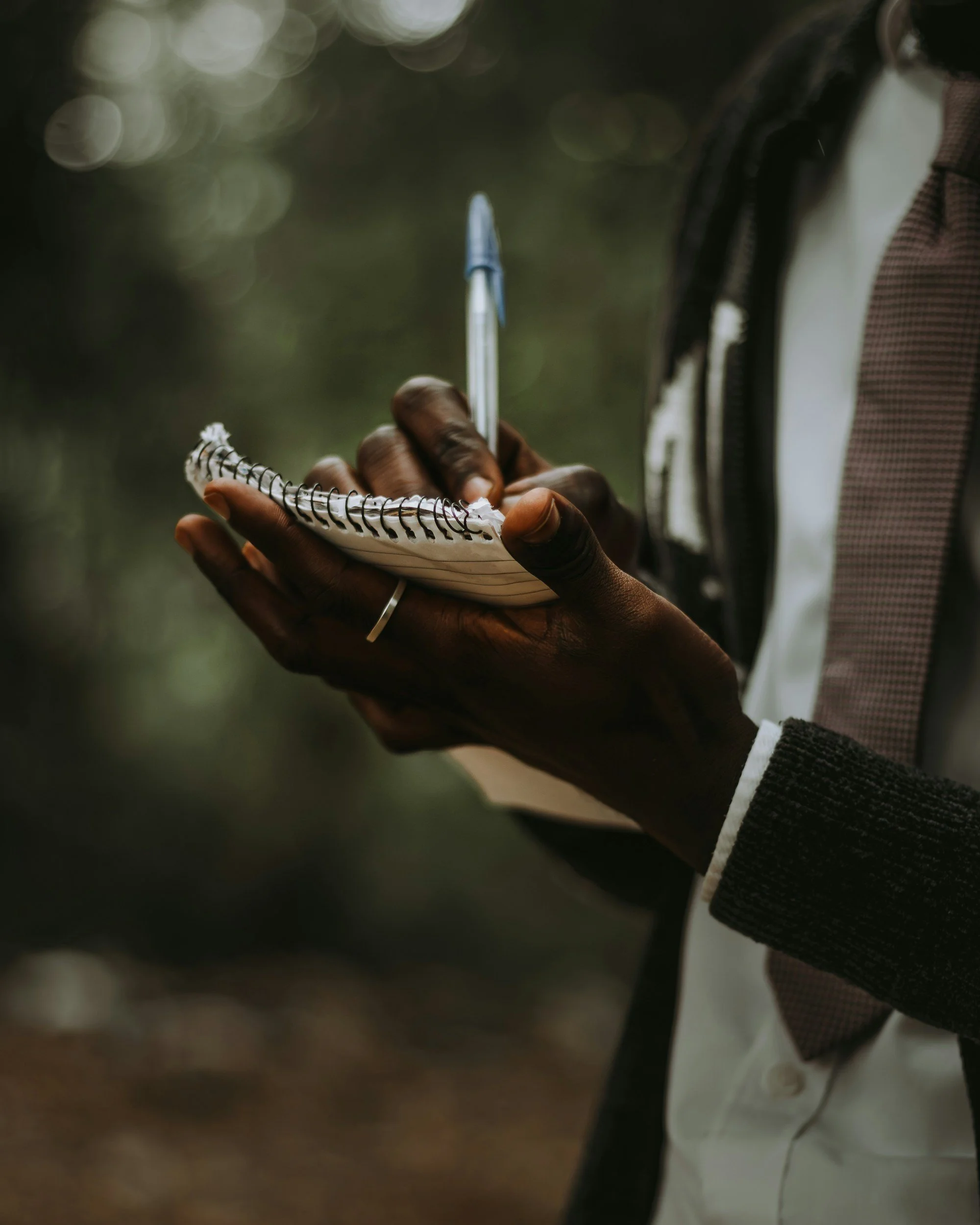Close-up of a person's hand holding a small spiral-bound notebook and a pen, with the person dressed in a checkered shirt, white turtleneck, and a dark jacket. The background is blurred with circular bokeh lights.