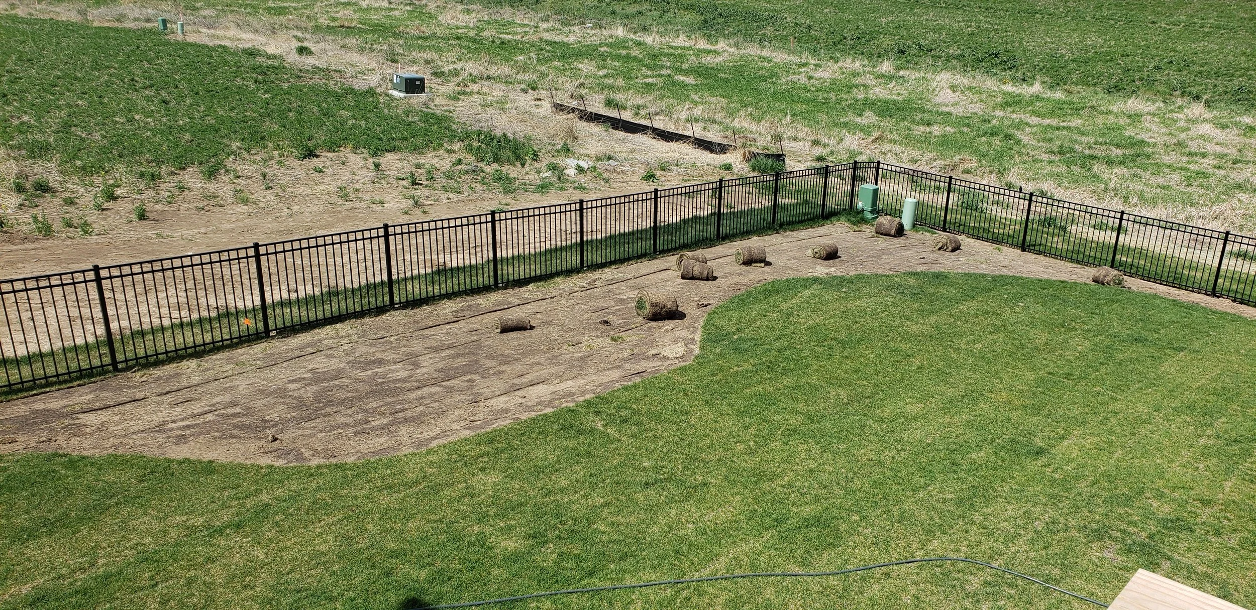 A fenced backyard with a grassy lawn, stone pathway, and scattered logs. There is a grassy field with some plants in the background.