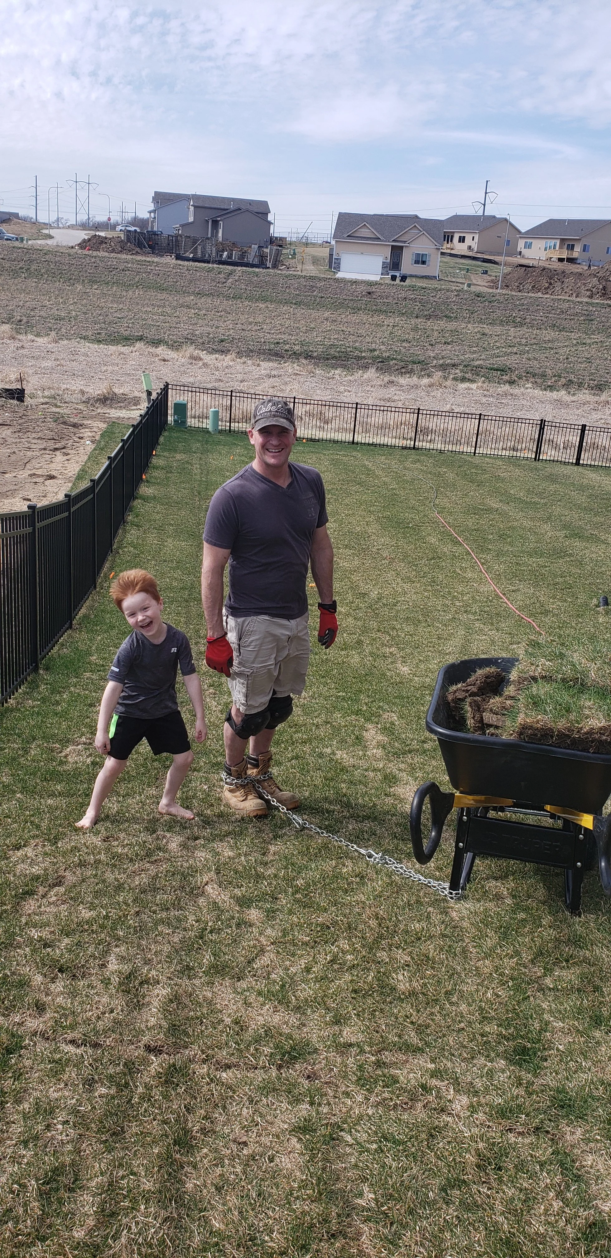 A man and a young boy standing in a backyard, smiling. The man is wearing work gloves, knee pads, boots, and has a chain attached to a wheelbarrow filled with sod or soil. The boy is barefoot and wearing a dark t-shirt and shorts. The yard is fenced, with houses and a hilly landscape in the background.
