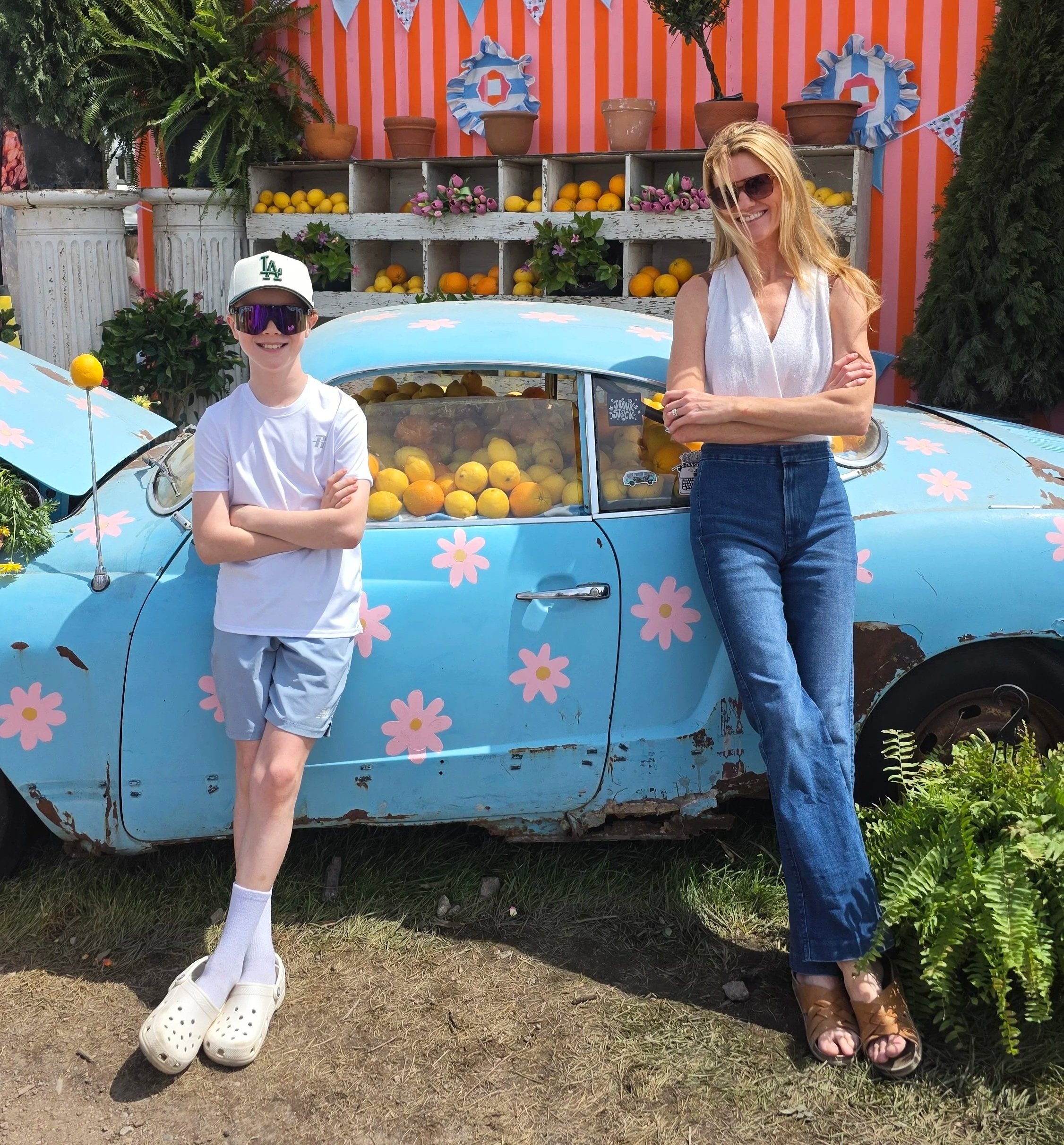 Two children standing in front of a vintage car decorated with pink flowers and yellow lemons, with potted plants and orange-striped background. The girl on the right is wearing sunglasses and crossing her arms, and the boy on the left is wearing sunglasses, a white cap, white t-shirt, and Crocs.