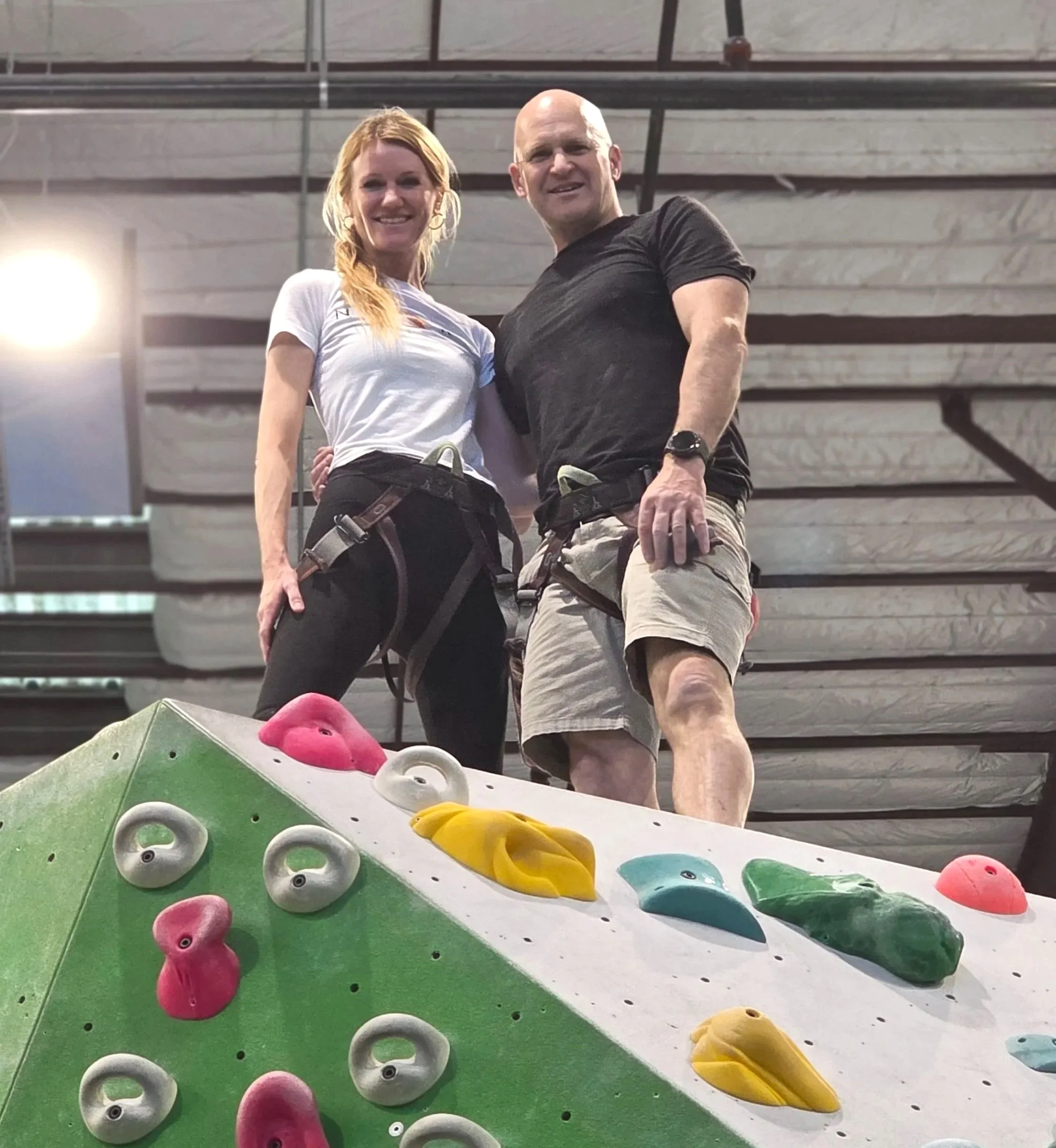 Indoor climbing gym with colorful climbing holds on a green wall and two climbers wearing harnesses ready to climb.