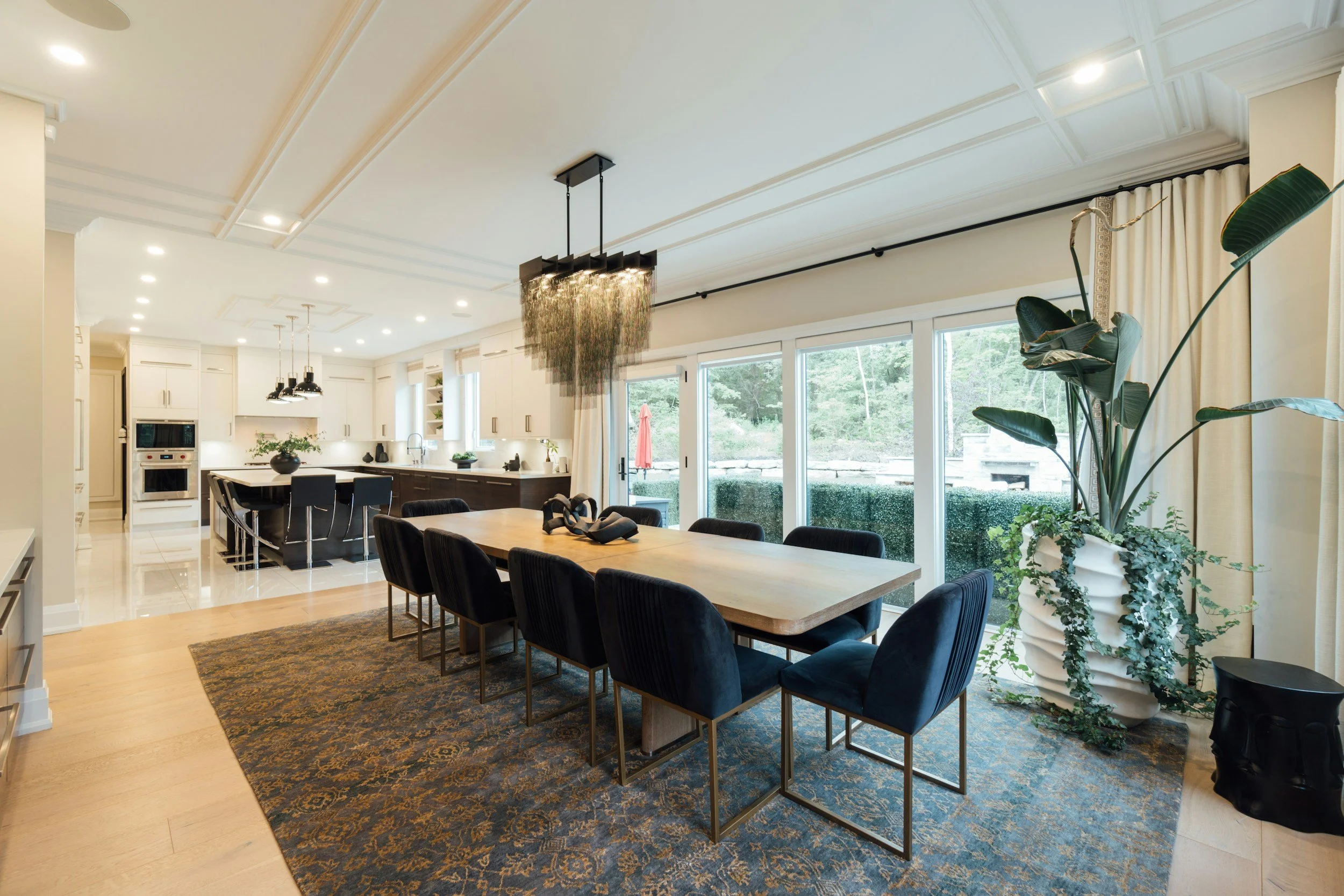 Open-concept kitchen and dining area with a large wooden dining table, black chairs, a kitchen island, white cabinets, large windows with curtains, and houseplants.