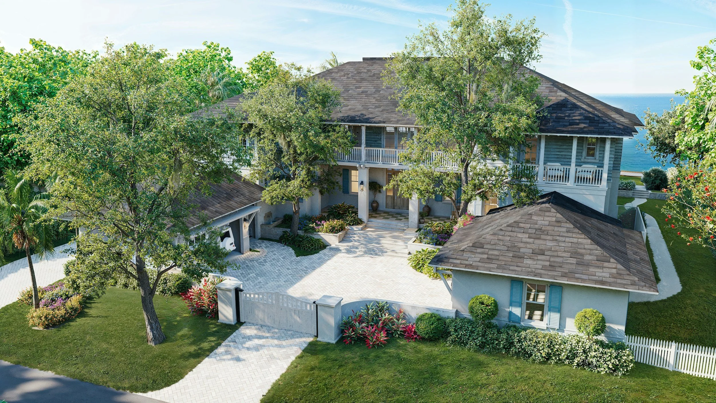A large, elegant house with a gray shingled roof, surrounded by lush greenery and colorful flowers. The house features a balcony, a driveway, and is near the ocean in the background.