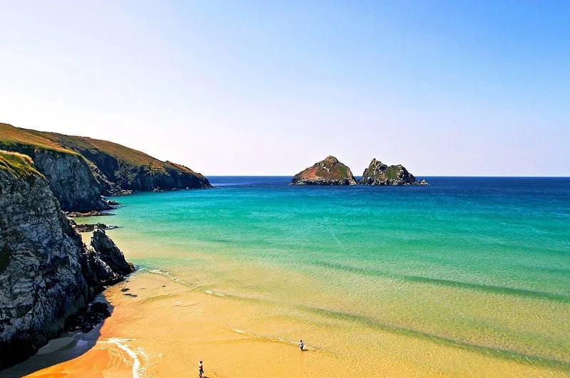 A scenic beach with yellow sand, green waters, rocky cliffs on the left, and two small islands in the distance under a clear blue sky.