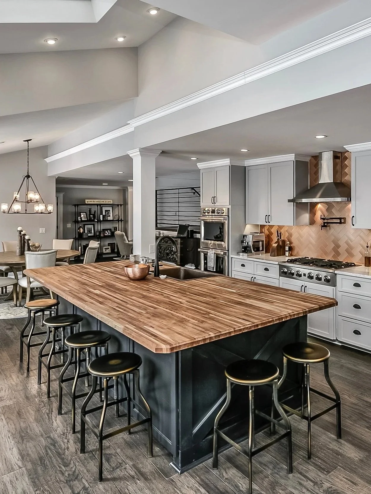 Modern remodeled kitchen with a large wooden island, black stools, white cabinets, copper backsplash, stainless steel appliances, and recessed lighting.